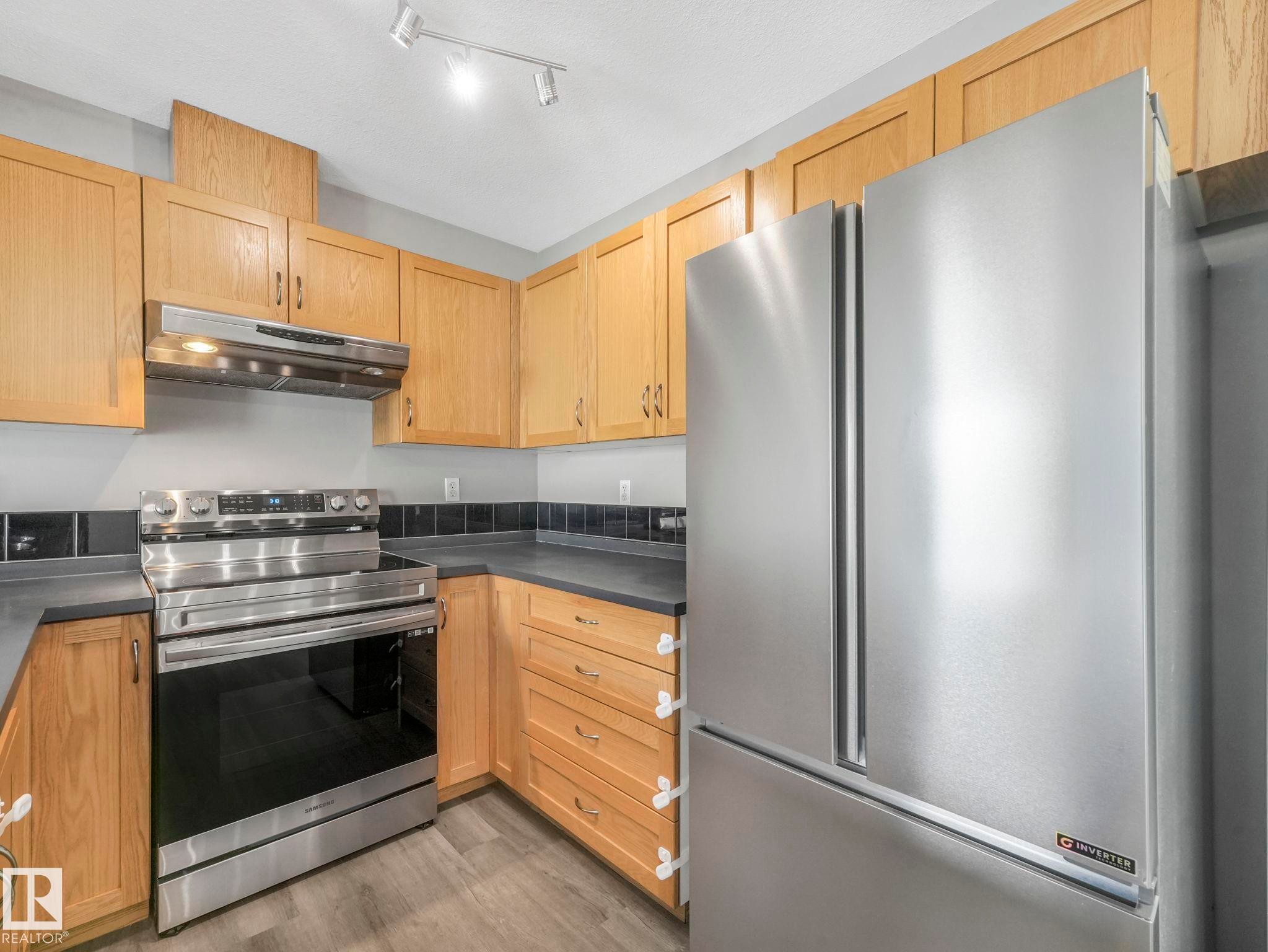 The kitchen features light wood cabinetry, dark countertops, a stainless steel range with an overhead vent, and a stainless steel French door refrigerator - 175 150 Edwards Drive, Edmonton, AB - Indoor Photo Showing Kitchen