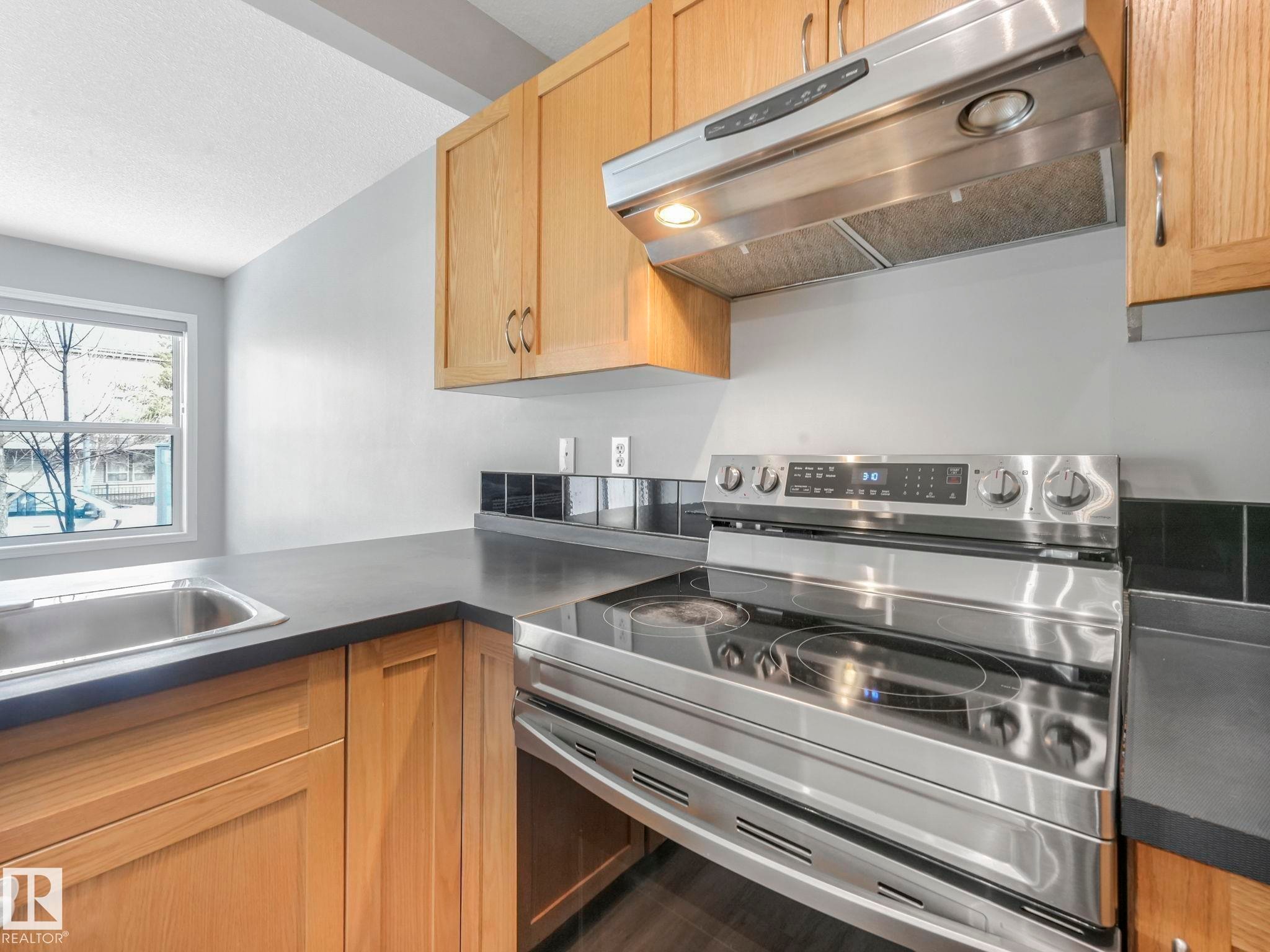 The kitchen features light wood cabinetry, dark countertops, a stainless steel range with a matching range hood, and a stainless steel sink with a window above it - 175 150 Edwards Drive, Edmonton, AB - Indoor Photo Showing Kitchen