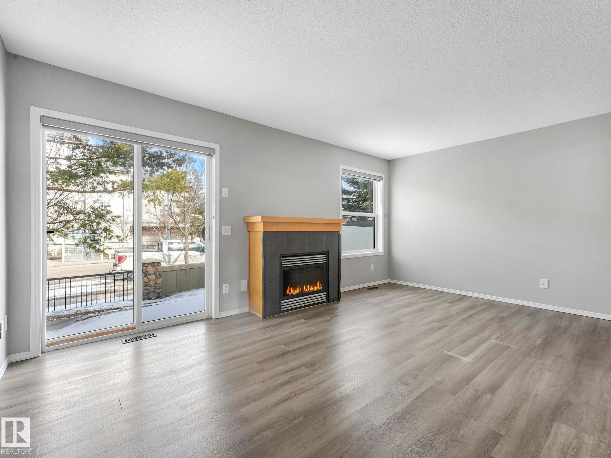 The living area features light grey walls, grey wood-style flooring, a modern fireplace with a light wood mantel, and large sliding glass doors providing natural light - 175 150 Edwards Drive, Edmonton, AB - Indoor Photo Showing Living Room With Fireplace