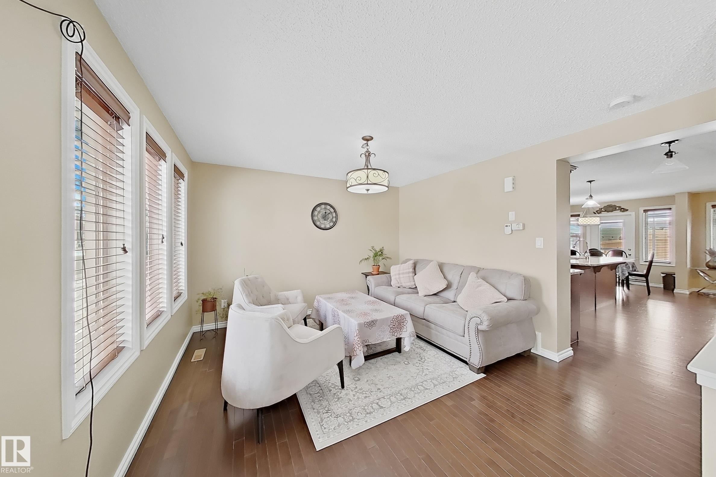 Living area featuring wood-finish flooring, light tan wall color, a decorative ceiling fixture, and multiple windows with horizontal blinds - 3609 Weidle Bend, Edmonton, AB - Indoor Photo Showing Living Room