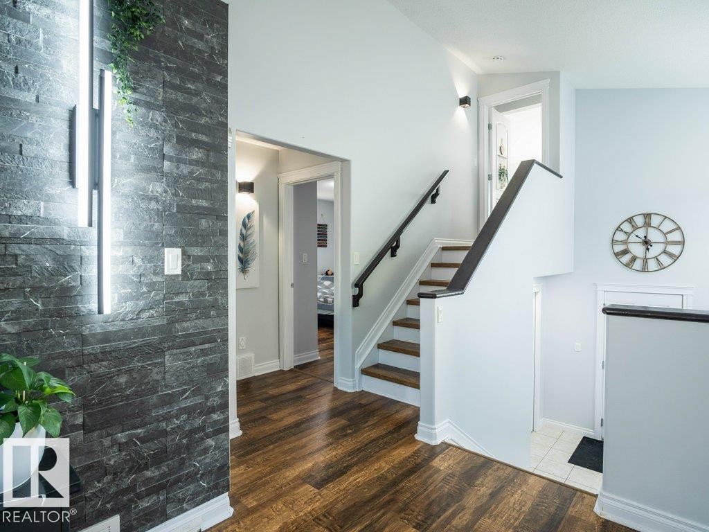 Interior entryway featuring a dark stacked stone accent wall with integrated vertical lighting, wood-finish flooring, a contemporary staircase with dark wood handrails, light-colored wall paint, and white trim throughout - 5701 Rue Eaglemont, Beaumont, AB - Indoor Photo Showing Other Room