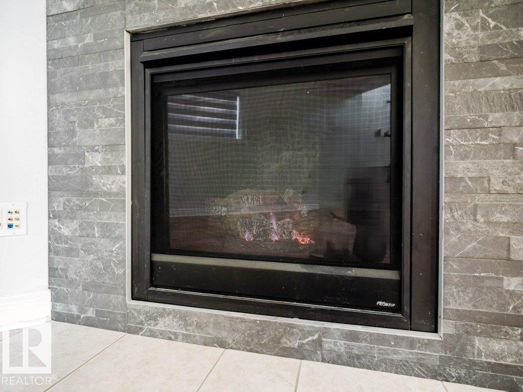Modern fireplace featuring a black metal frame, gray stacked stone surround, and light tile flooring - 5701 Rue Eaglemont, Beaumont, AB - Indoor Photo Showing Other Room