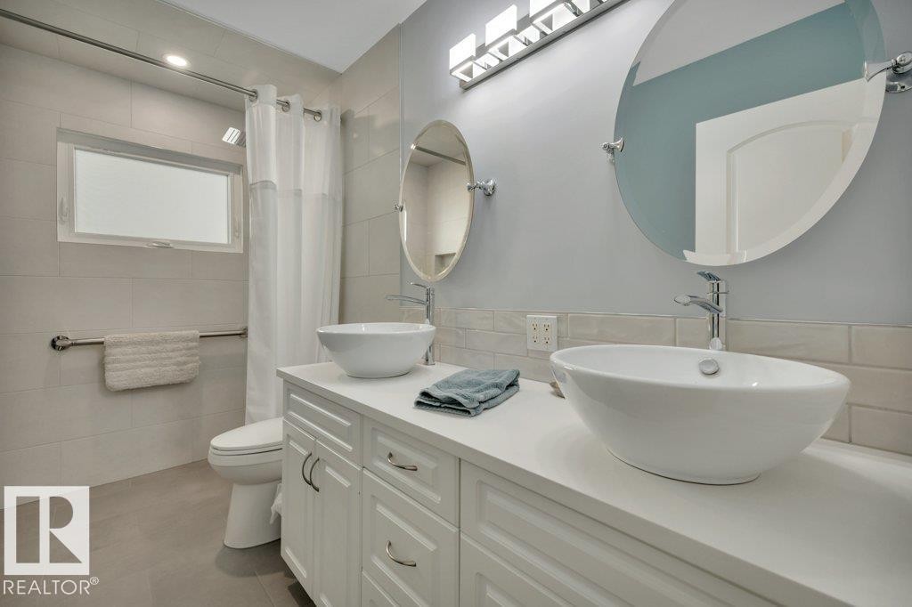 Dual vessel sinks with chrome faucets, white vanity cabinetry with chrome hardware, oval frameless mirrors, and a tiled shower surround with a window - 10633 St Gabriel School Road, Edmonton, AB - Indoor Photo Showing Bathroom