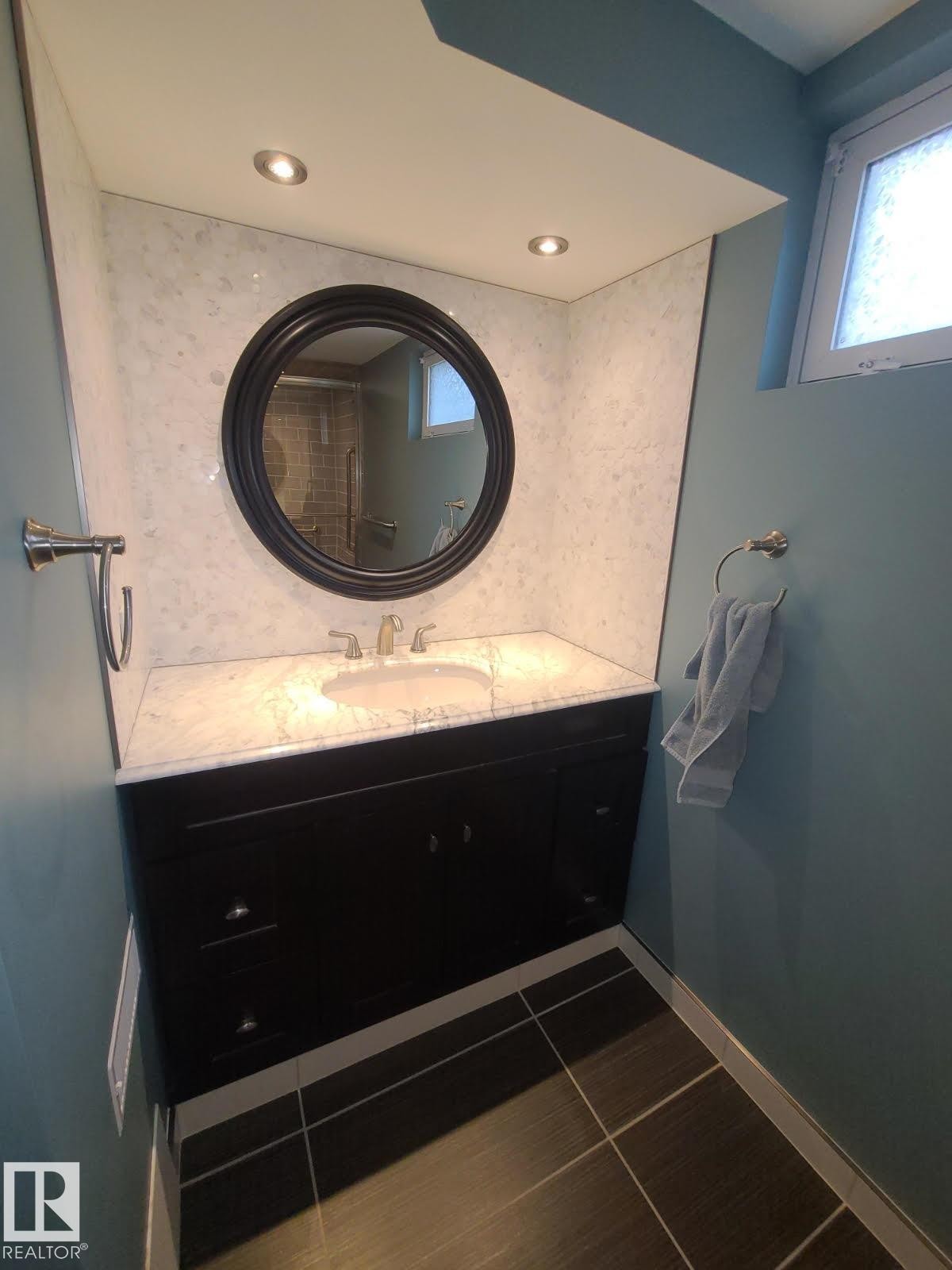 Vanity area featuring a dark wood-finish cabinet with an undermount sink, a stone countertop, a circular mirror, recessed lighting, and large format floor tiling - 10633 St Gabriel School Road, Edmonton, AB - Indoor Photo Showing Bathroom