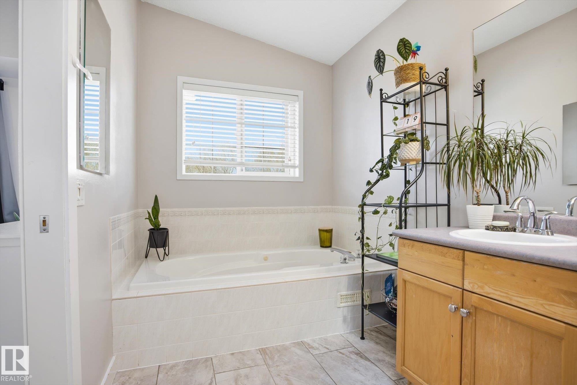 Soaking tub with a tiled surround, window with horizontal blinds, light-toned wood vanity, integrated sink, and chrome faucet - 2251 Brennan Court, Edmonton, AB - Indoor Photo Showing Bathroom