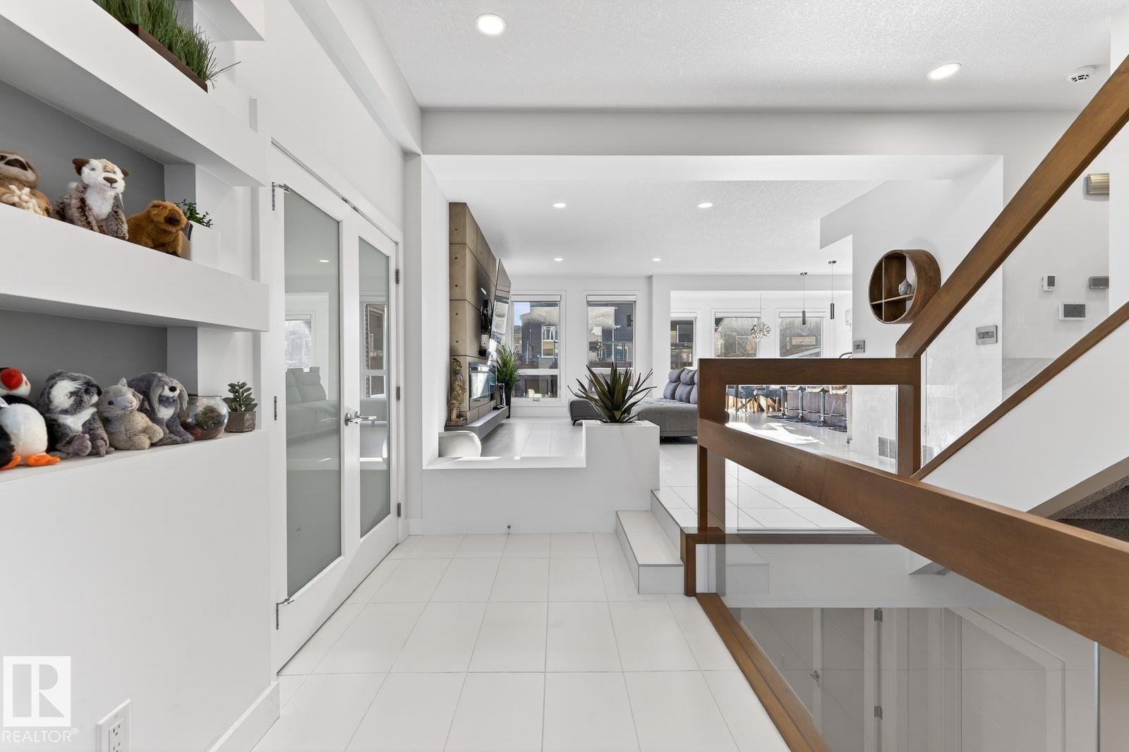 Contemporary open-concept interior featuring a glass and wood-finish staircase, white tile flooring, built-in shelving, recessed lighting, and a double-door entry with frosted glass - 793 Windermere Wynd, Edmonton, AB - Indoor Photo Showing Other Room