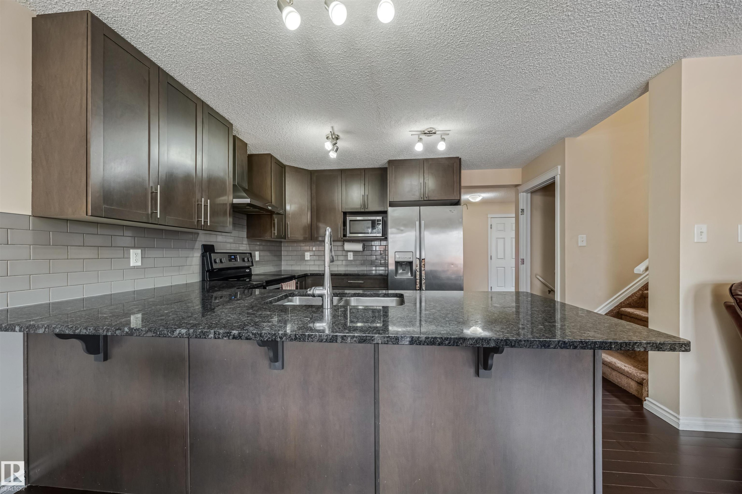 Kitchen featuring dark wood-finish cabinetry, stainless steel appliances, a subway tile backsplash, and a large central island with a dark stone-finish countertop and undermount sink - 2314 22 Avenue, Edmonton, AB - Indoor Photo Showing Kitchen With Upgraded Kitchen