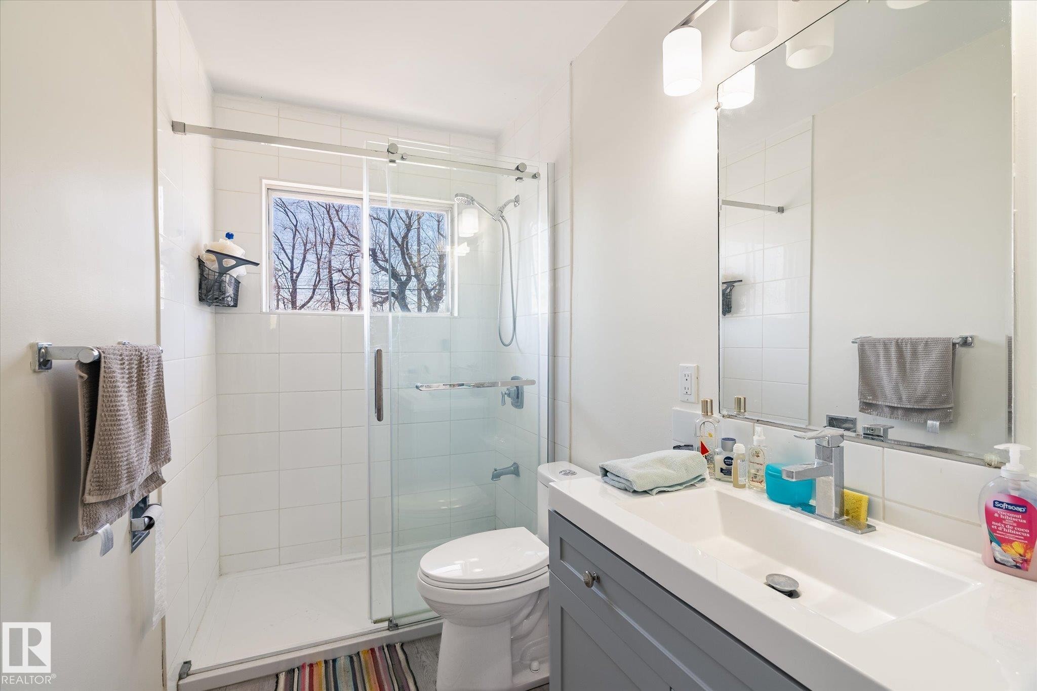 Modern bathroom featuring a glass-enclosed shower with white subway tile, a window, a single vanity with a white countertop, and an integrated sink with a chrome faucet - 10531 128 Avenue, Edmonton, AB - Indoor Photo Showing Bathroom