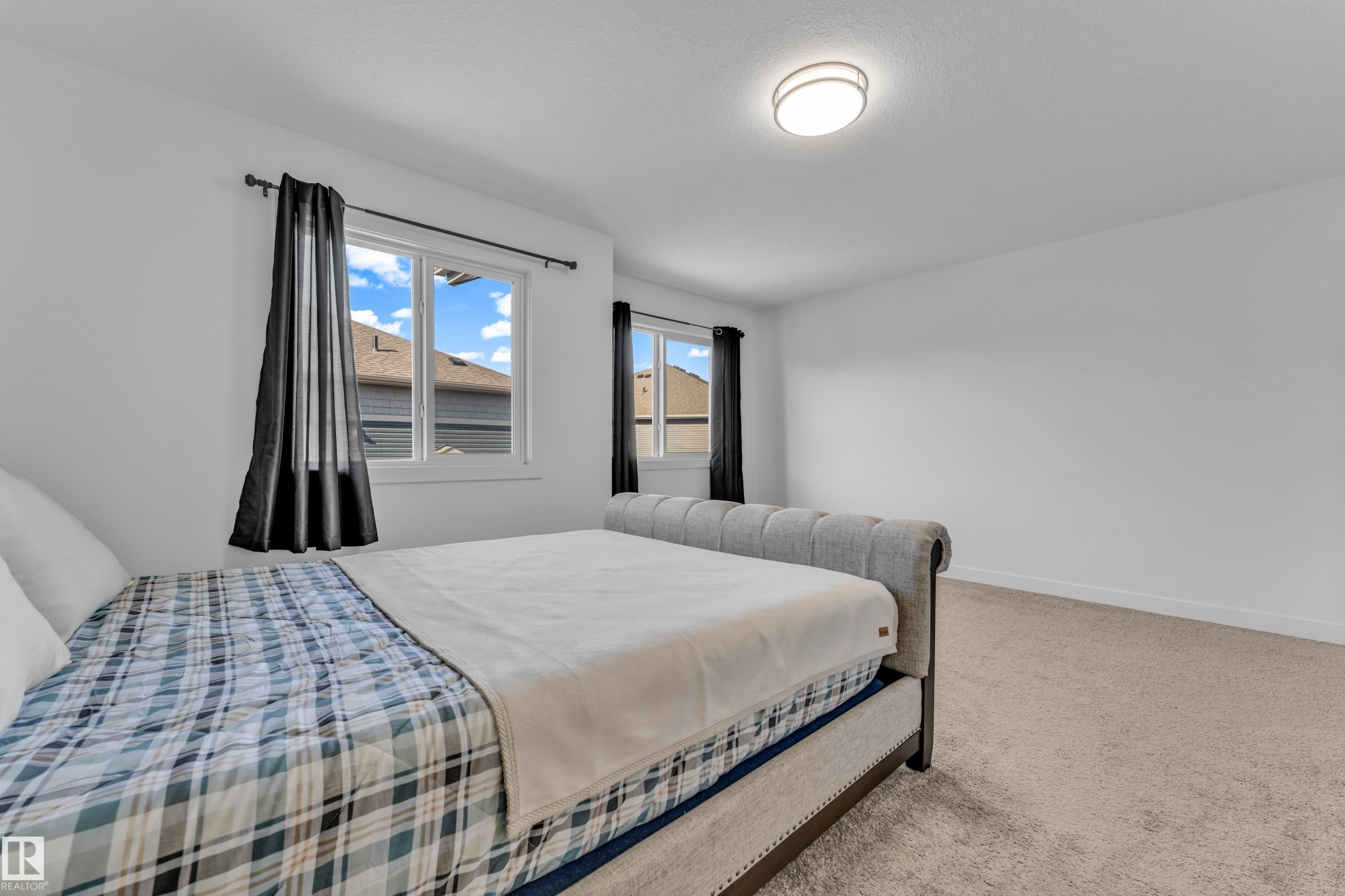 Bright room featuring two windows with dark drapes, light carpet flooring, and a flush-mount ceiling light fixture - 41 603 Orchards Boulevard, Edmonton, AB - Indoor Photo Showing Bedroom