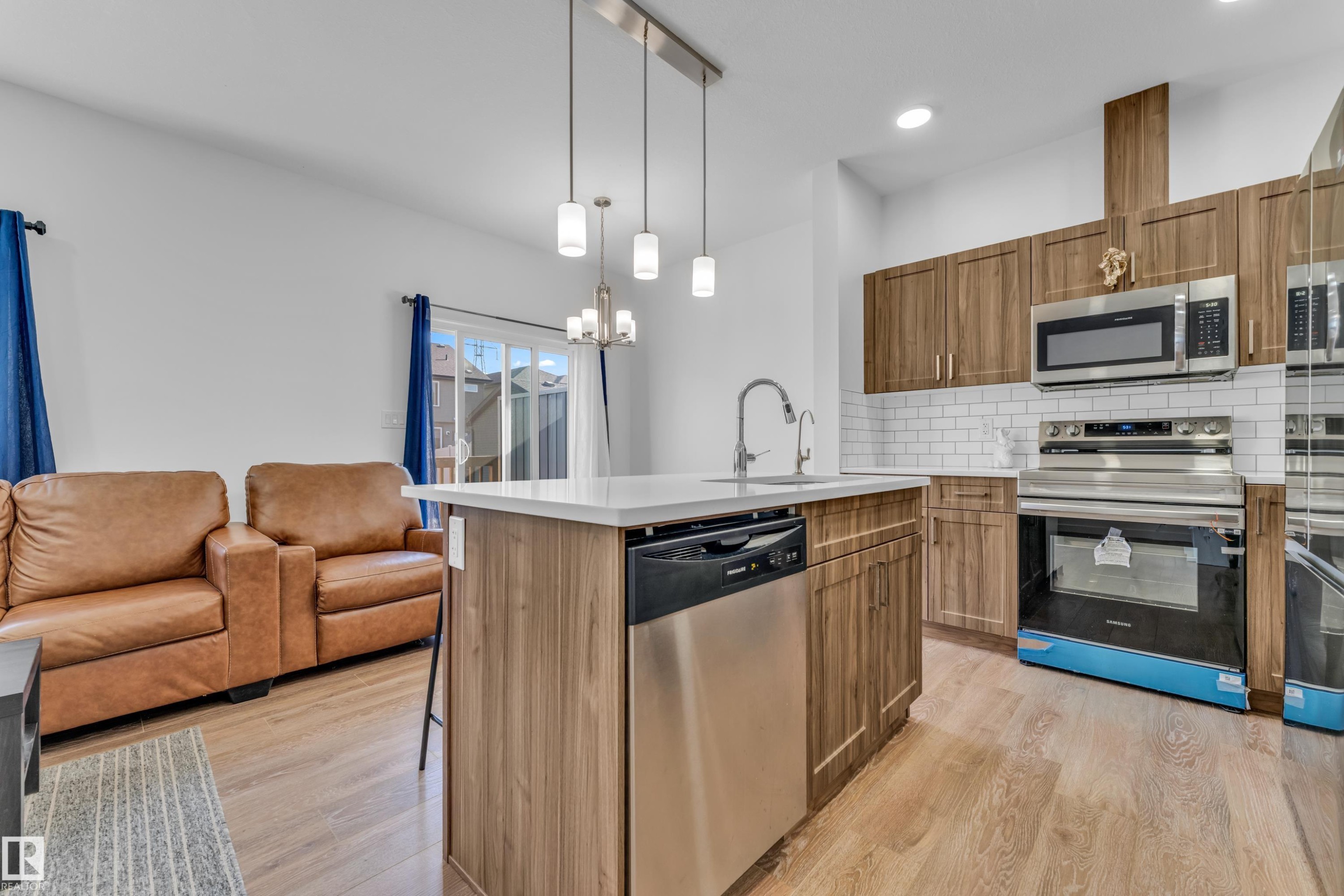 Kitchen island with light-toned countertop, stainless steel appliances, wood-finish cabinetry, subway tile backsplash, and light wood-finish flooring - 41 603 Orchards Boulevard, Edmonton, AB - Indoor Photo Showing Kitchen