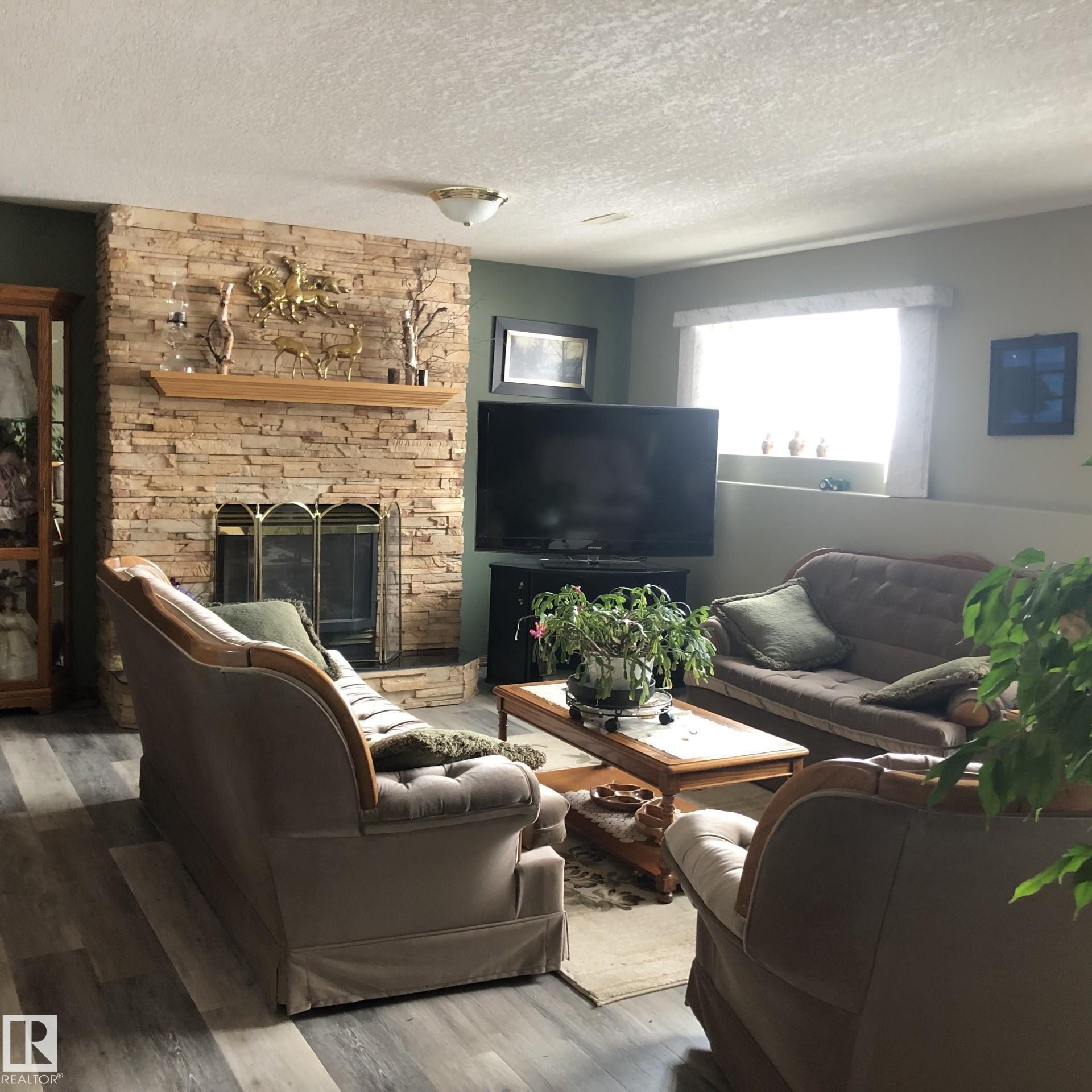 Living area featuring a floor-to-ceiling stacked stone fireplace with a wood mantle and brass-toned screen, wood-finish flooring, recessed window, and wall-mounted lighting - 123064 Hwy 45, Rural Two Hills County, AB