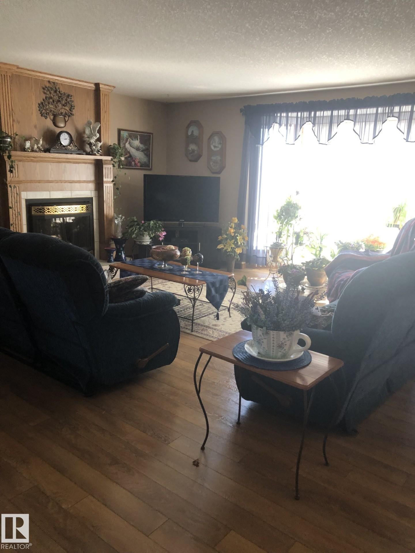 Living room featuring wood-finish flooring, a fireplace with a stone surround and wood mantle, and a large window with valance - 123064 Hwy 45, Rural Two Hills County, AB
