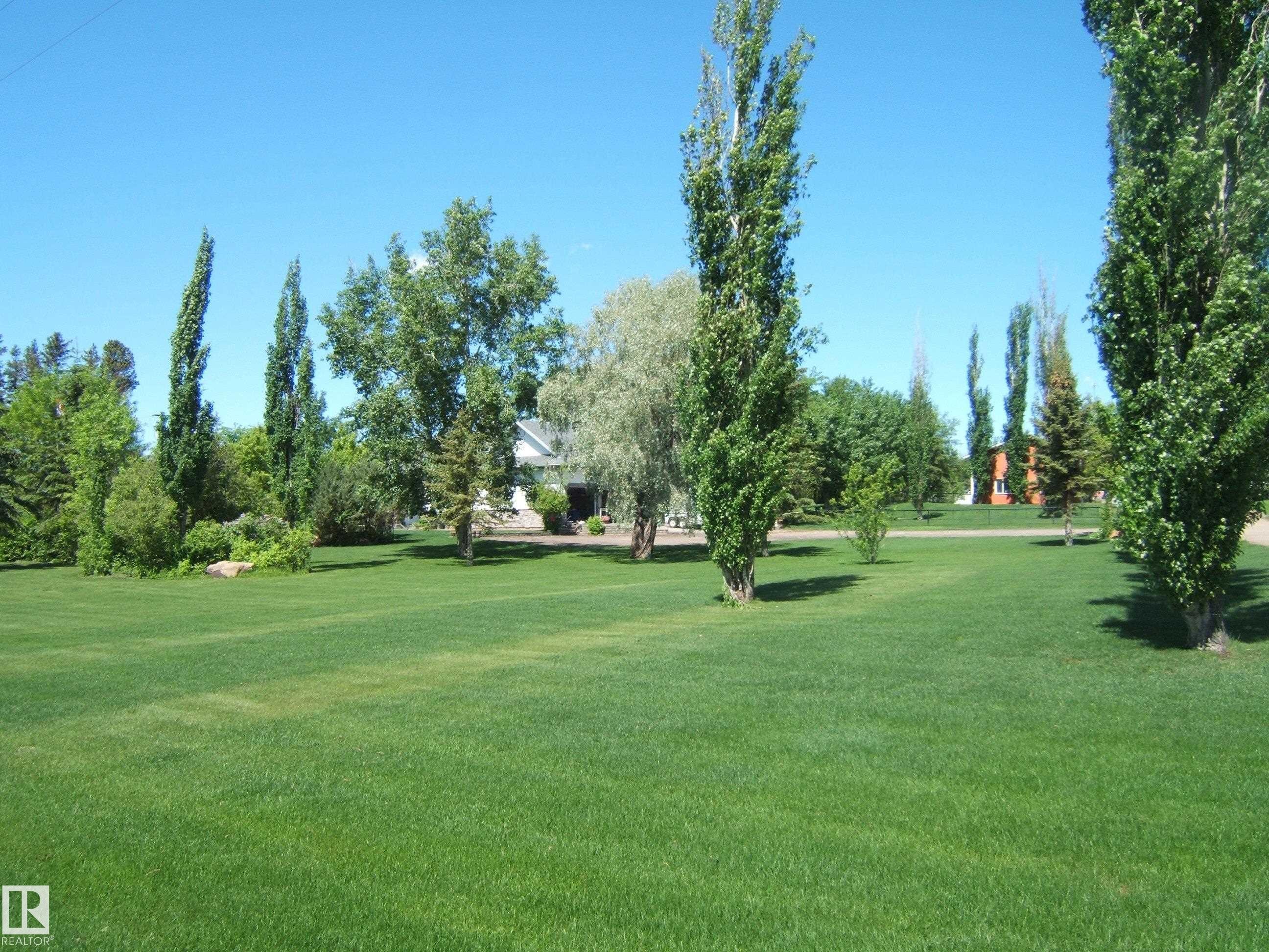 Expansive green lawn framed by mature trees, featuring a distant white residence and a separate orange structure - 123064 Hwy 45, Rural Two Hills County, AB