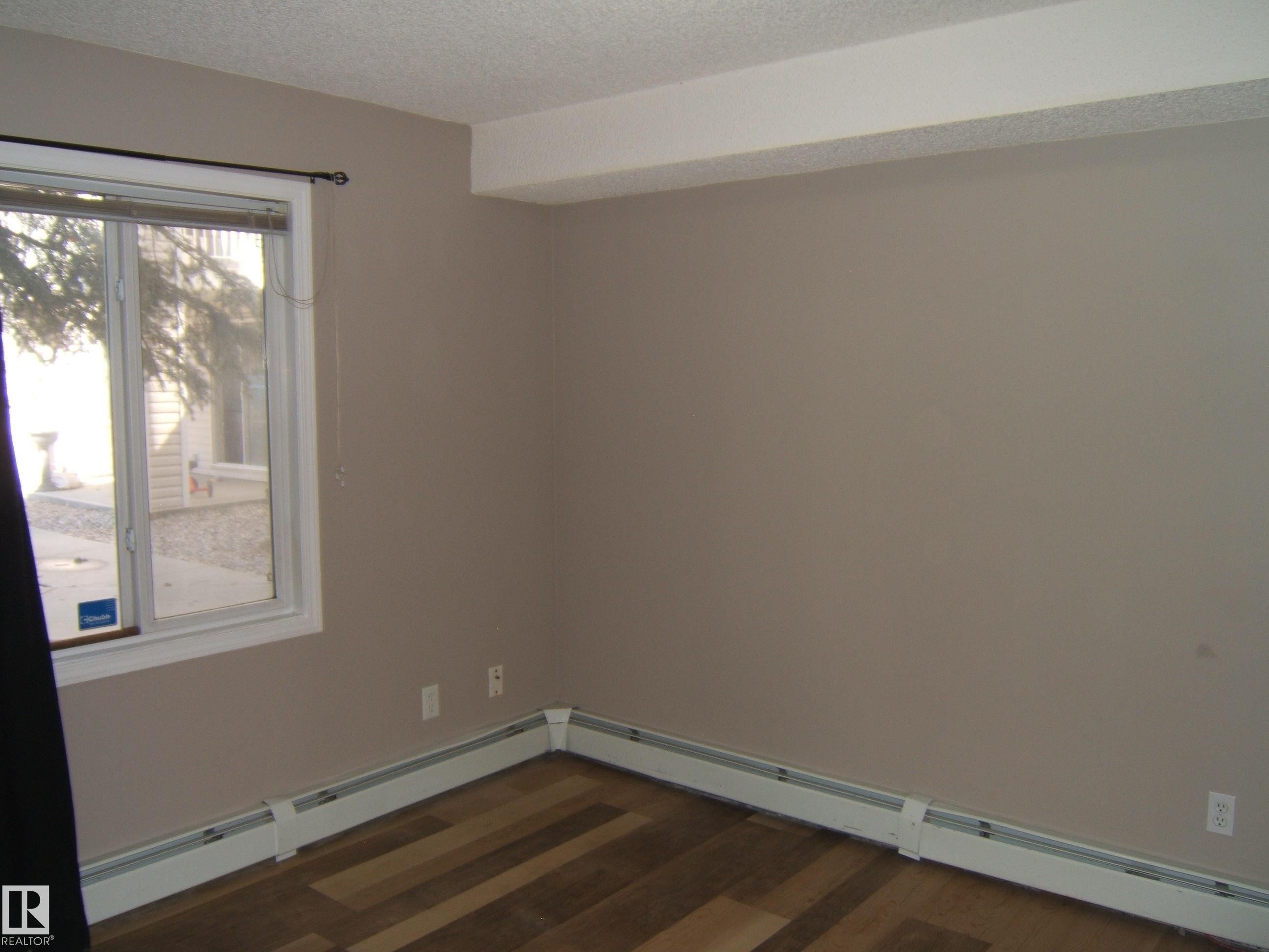 Room featuring wood-finish flooring, a window with white trim, and baseboard heating - 105 11214 80 Street, Edmonton, AB - Indoor Photo Showing Other Room