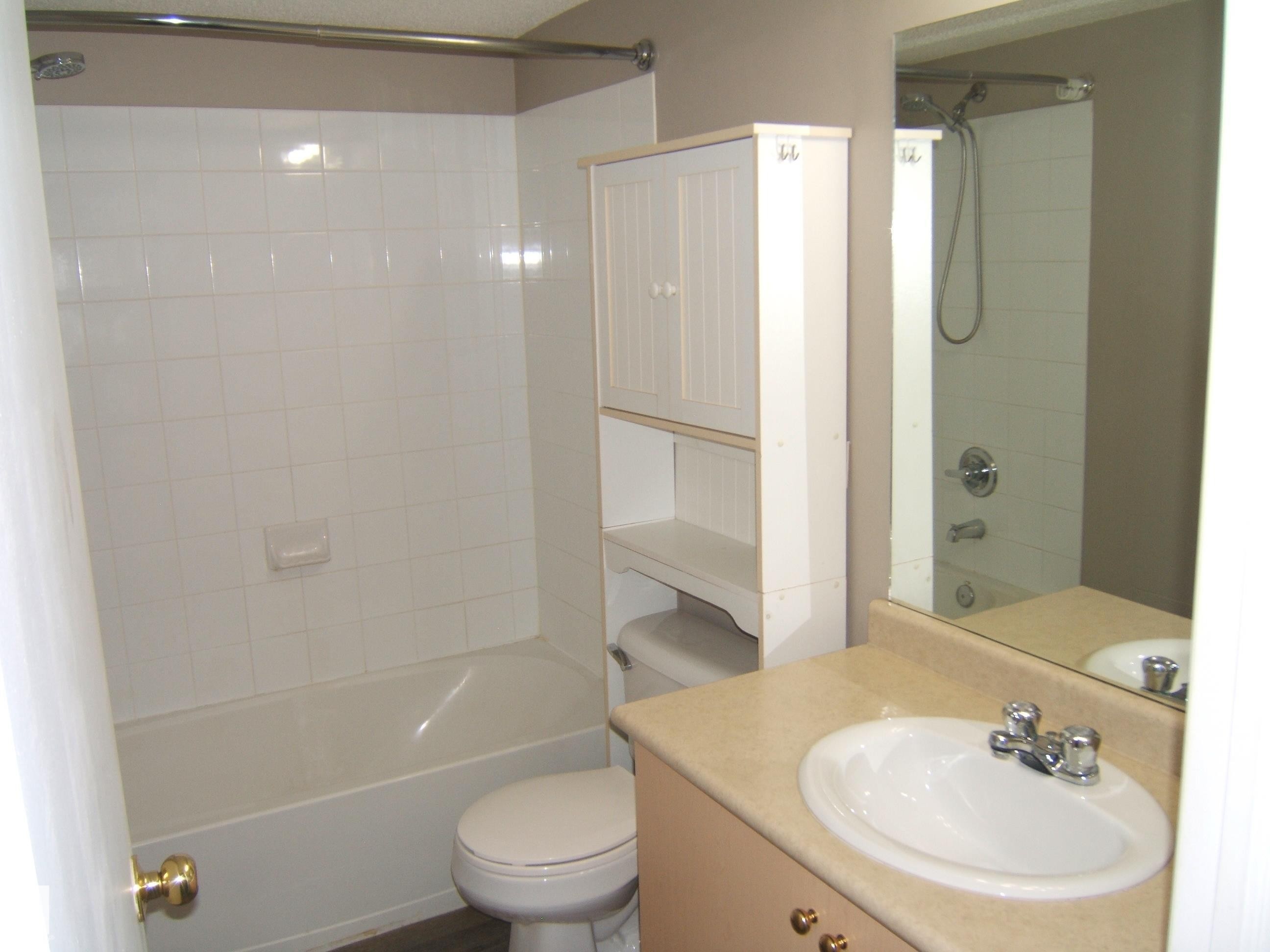 Bathroom featuring a white tiled bath and shower combination, a white commode, and a light-toned vanity with an integrated sink and chrome faucet - 105 11214 80 Street, Edmonton, AB - Indoor Photo Showing Bathroom