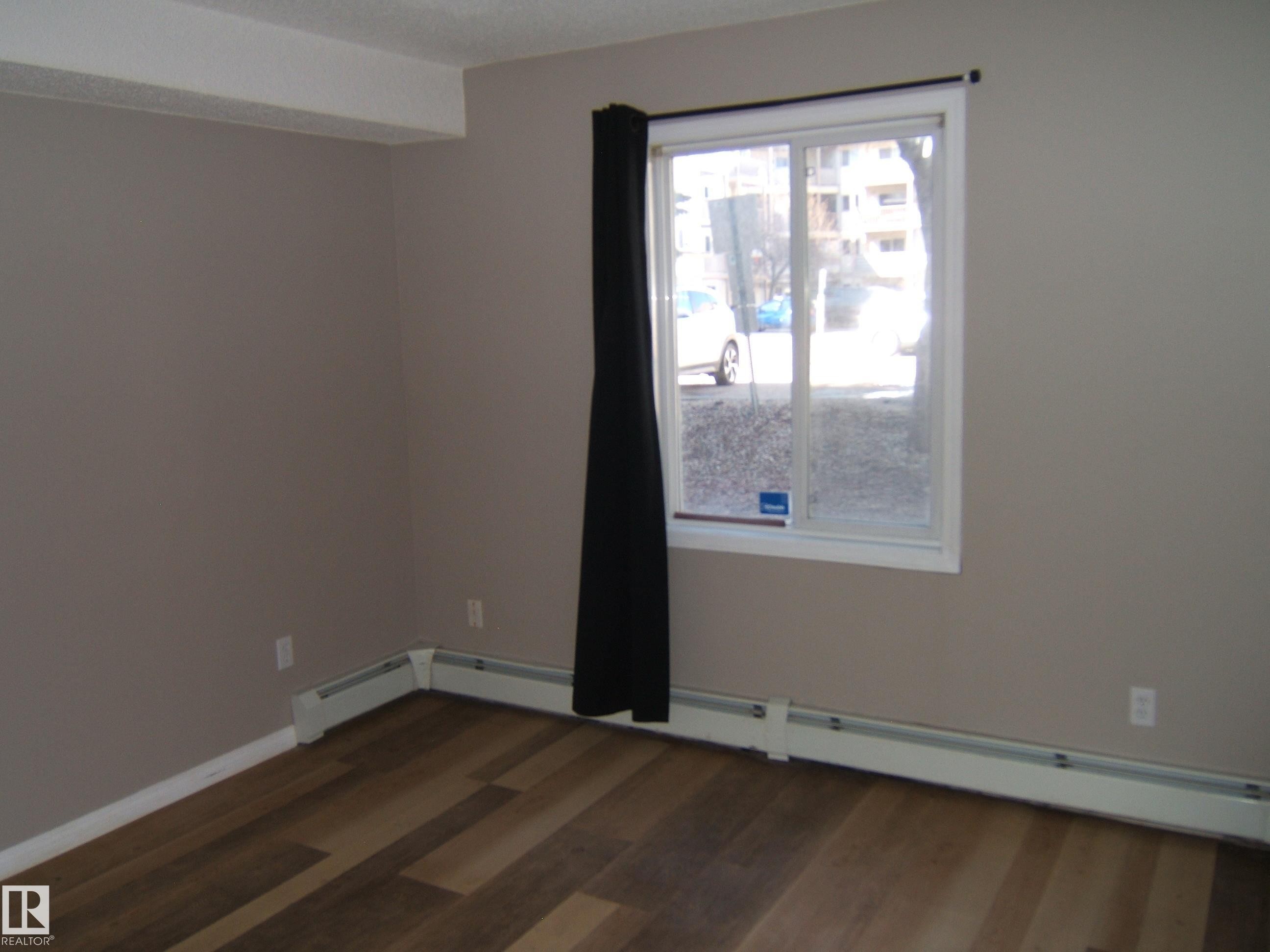 Neutral tone walls with wood-finish flooring, white baseboards, and a window featuring a white frame - 105 11214 80 Street, Edmonton, AB - Indoor Photo Showing Other Room