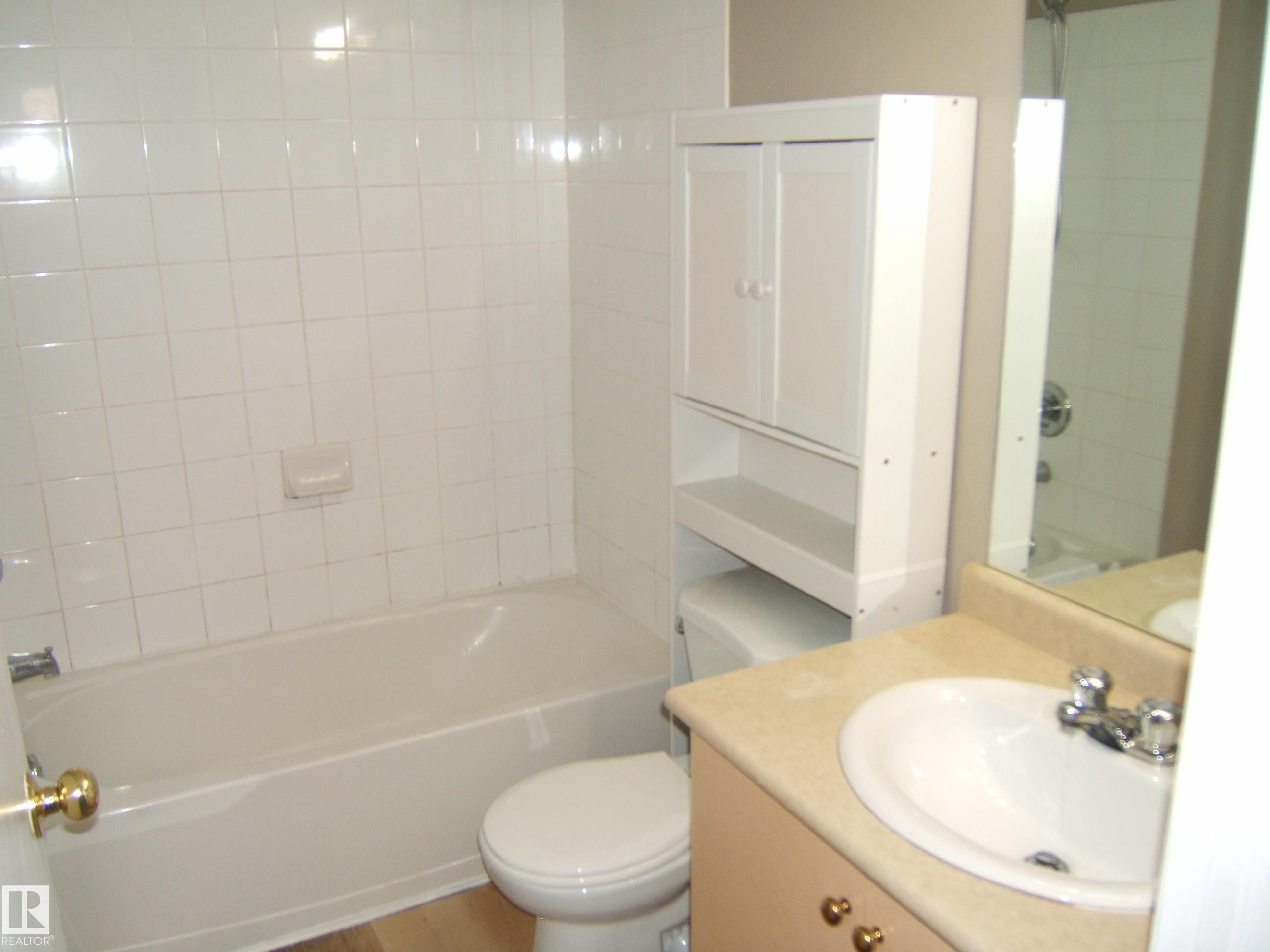 Bathroom with a white tiled tub surround, a built-in white over-toilet cabinet, and a vanity with an integrated sink and mirror - 105 11214 80 Street, Edmonton, AB - Indoor Photo Showing Bathroom