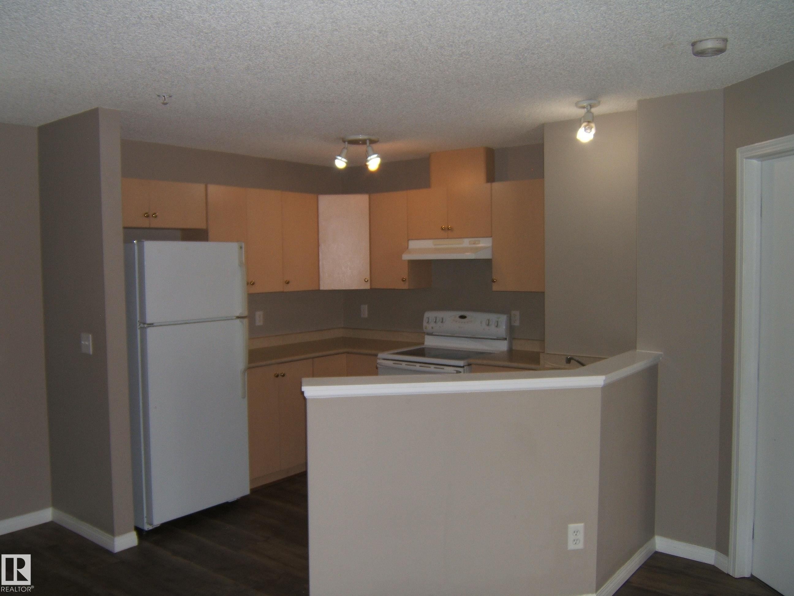 Kitchen featuring wood-finish flooring, light wood cabinetry, white appliances, and a breakfast bar peninsula - 105 11214 80 Street, Edmonton, AB - Indoor Photo Showing Kitchen