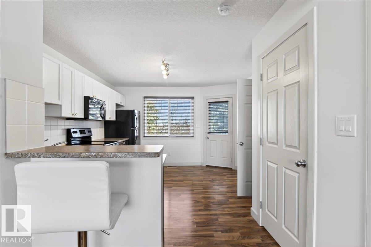 Kitchen featuring white cabinetry, black appliances, a breakfast bar, wood-finish flooring, and a window with blinds - 18 6032 38 Avenue, Edmonton, AB - Indoor Photo Showing Kitchen