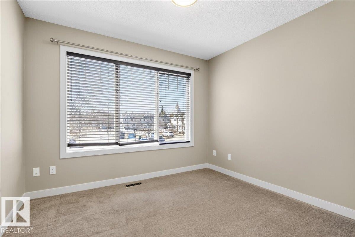 Neutral-toned room featuring a large window with blinds, carpeted flooring, and white baseboards - 18 6032 38 Avenue, Edmonton, AB - Indoor Photo Showing Other Room