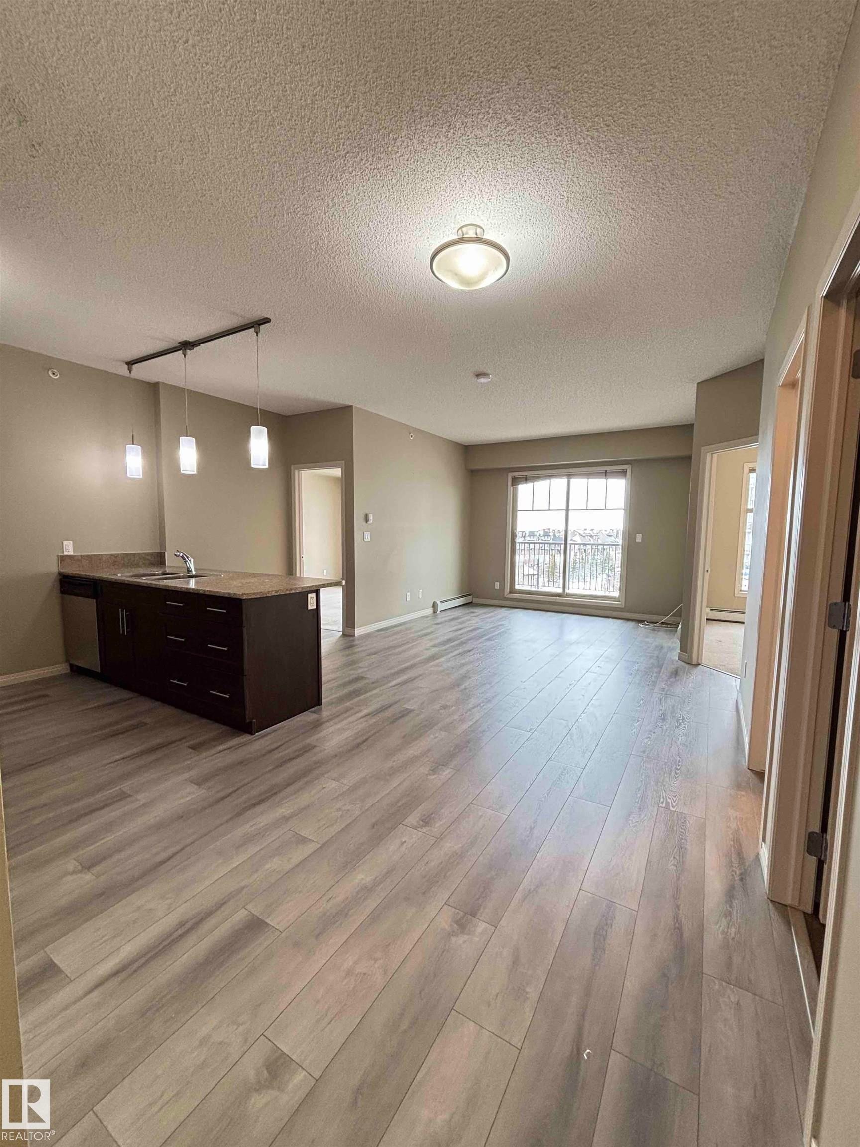 Open-concept living area featuring light wood-finish flooring, a kitchen island with dark cabinetry and a stone-finish countertop, a stainless steel dishwasher, and track pendant lighting - 408 111 Ambelside Drive Sw, Edmonton, AB - Indoor