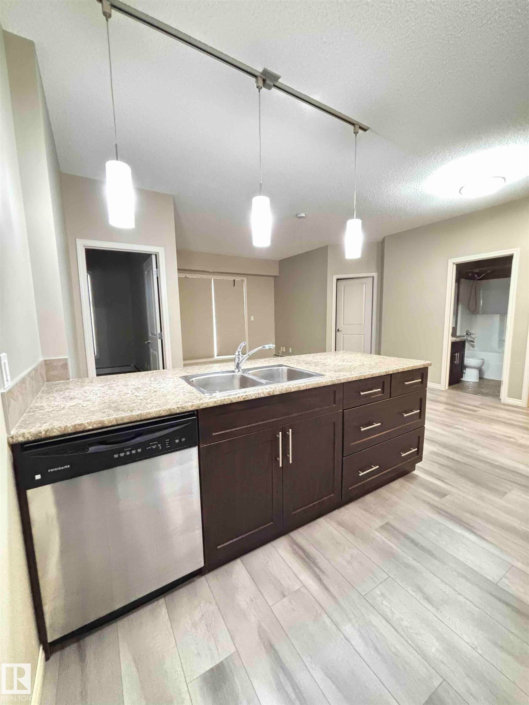 Kitchen island featuring a double basin stainless steel sink, granite countertop, dark wood cabinetry with chrome hardware, and an integrated stainless steel dishwasher - 408 111 Ambelside Drive Sw, Edmonton, AB - Indoor Photo Showing Kitchen With Double Sink