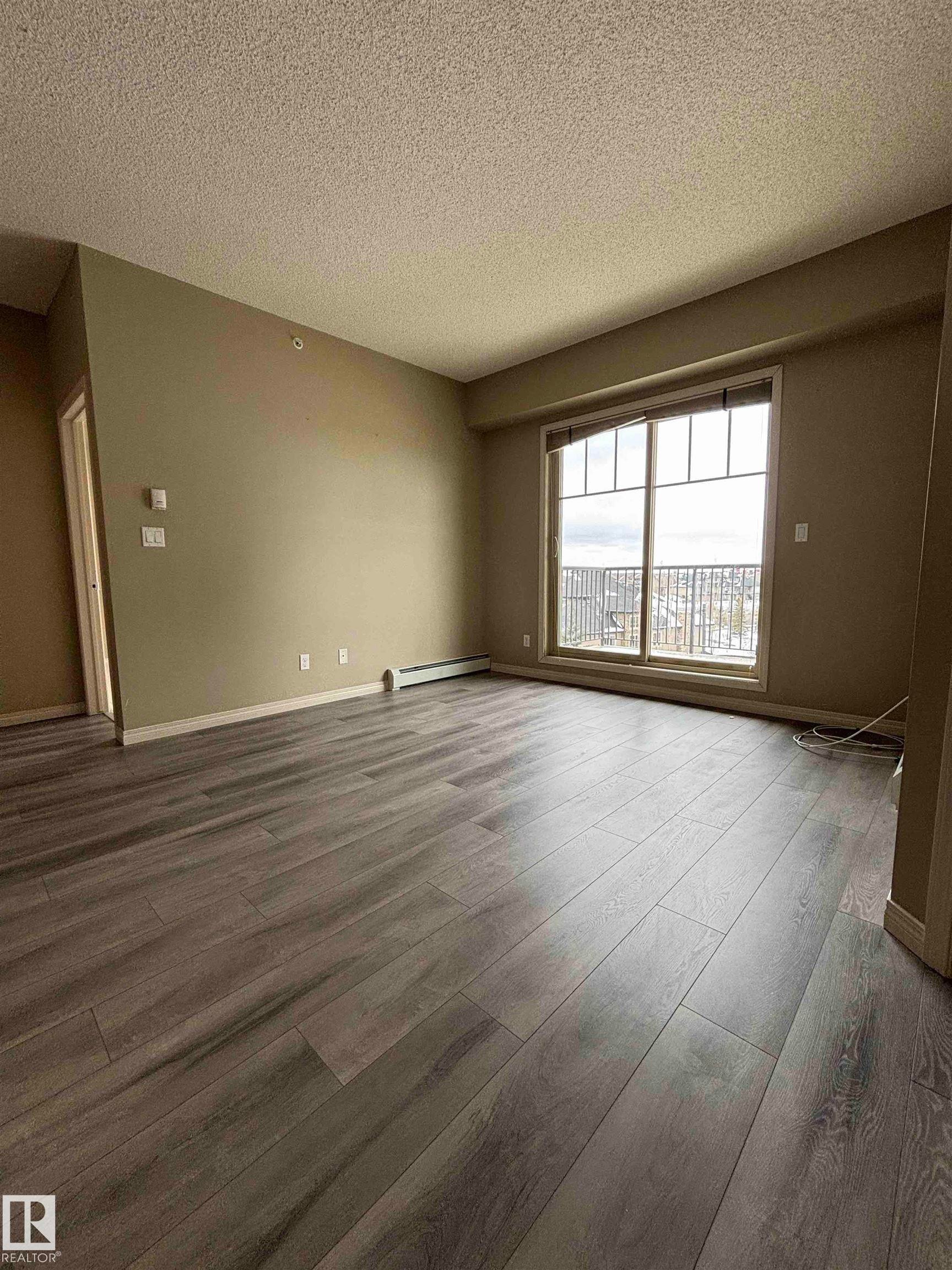 Spacious room featuring wood-finish flooring, a large window, neutral wall paint, and a textured ceiling - 408 111 Ambelside Drive Sw, Edmonton, AB - Indoor Photo Showing Other Room