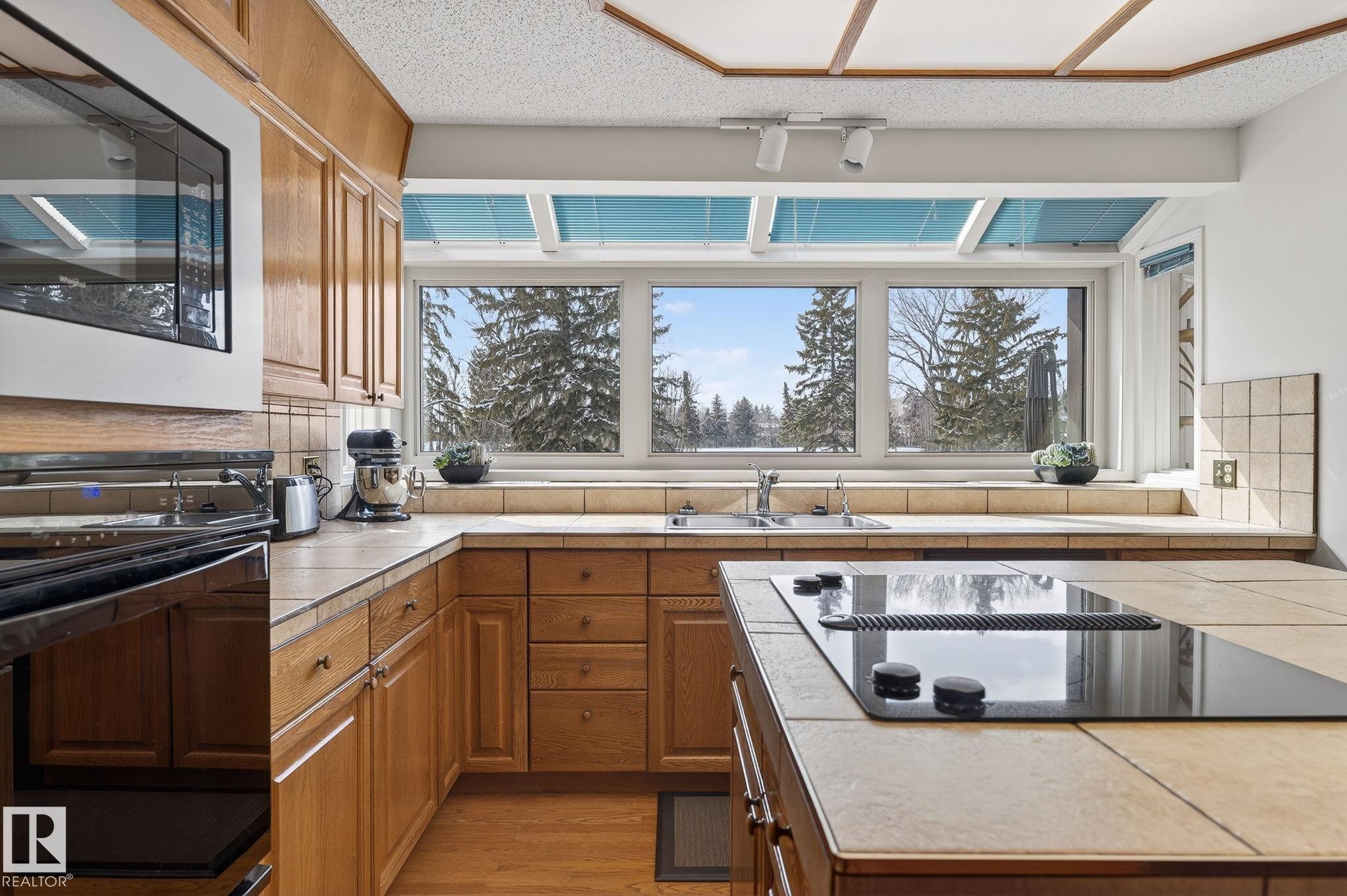 Kitchen featuring wood cabinetry, tile countertops, an integrated cooktop on the island, a built-in microwave, and a window expanse with upper transom panels - 848 Wheeler Road W, Edmonton, AB - Indoor Photo Showing Kitchen With Double Sink