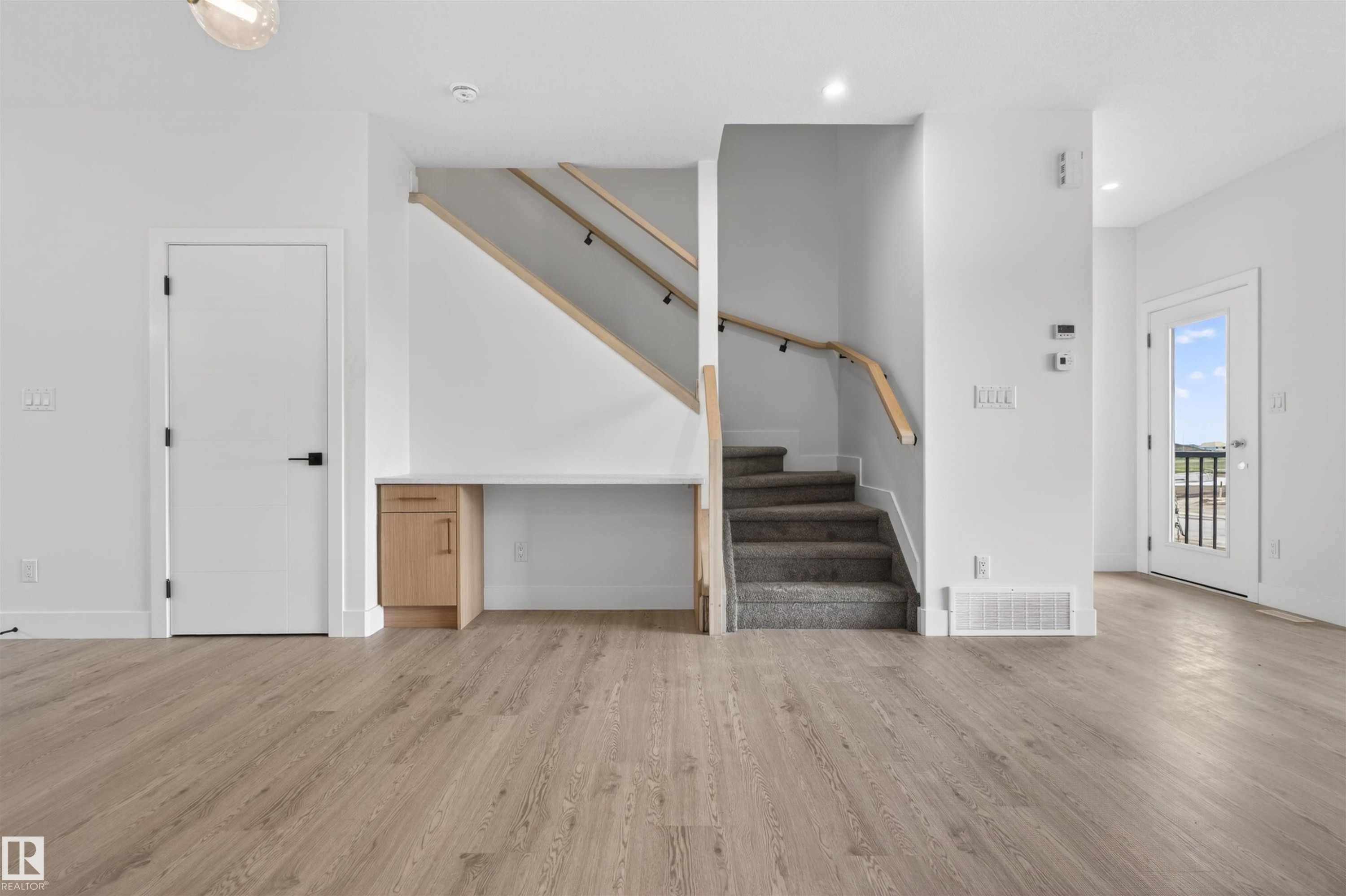 Open concept living area featuring wood-finish flooring, a built-in desk with natural wood-tone cabinetry, a staircase with carpeted treads and light wood handrails, white interior doors with black hardware, and an exterior door with a window - 45 River Birch, St. Albert, AB - Indoor Photo Showing Other Room