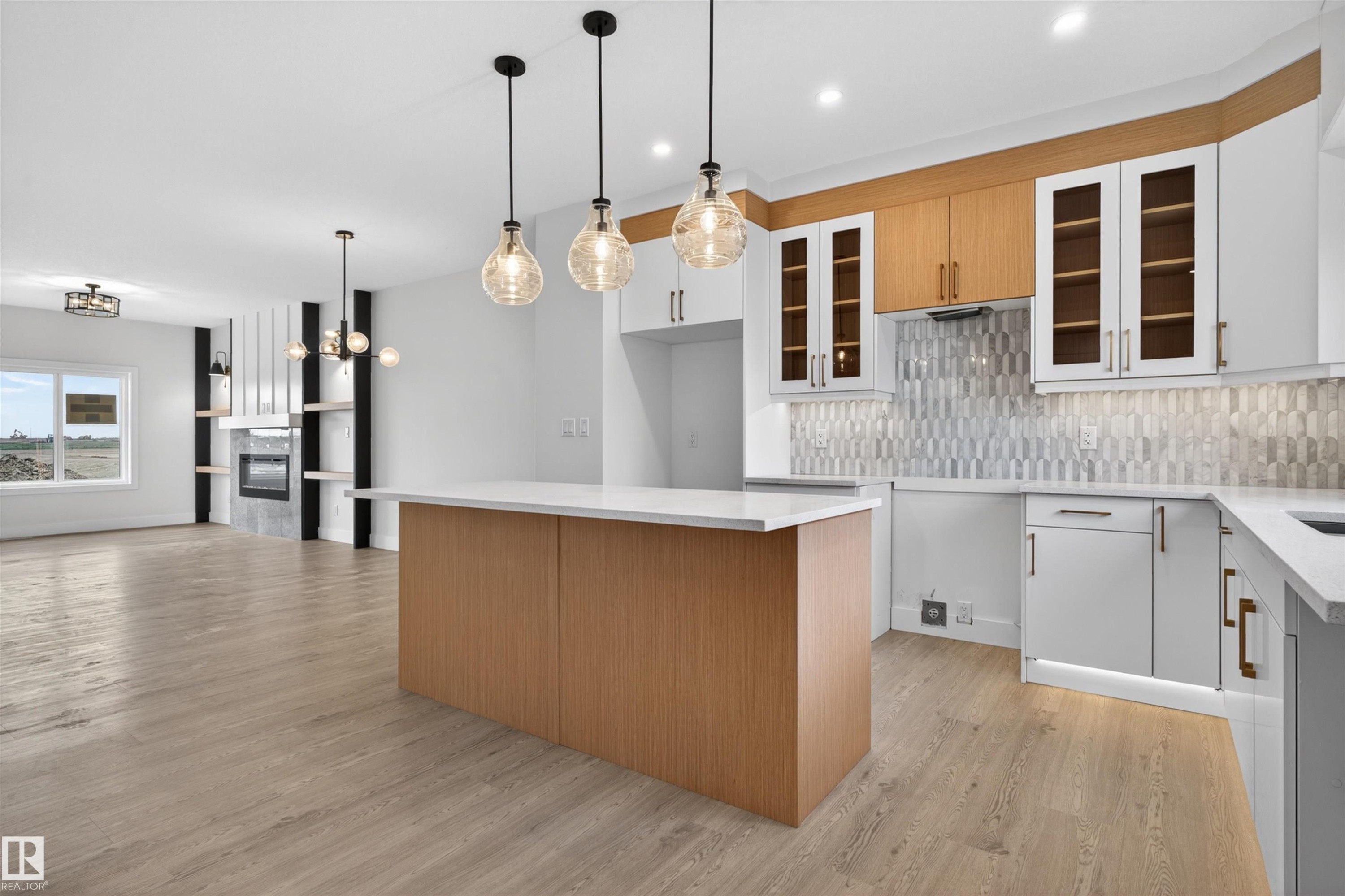 Spacious kitchen and living area featuring a large center island with a white countertop, two-tone cabinetry including clear-front upper cabinets, a geometric tile backsplash, and wood-finish flooring - 45 River Birch, St. Albert, AB - Indoor Photo Showing Kitchen With Upgraded Kitchen