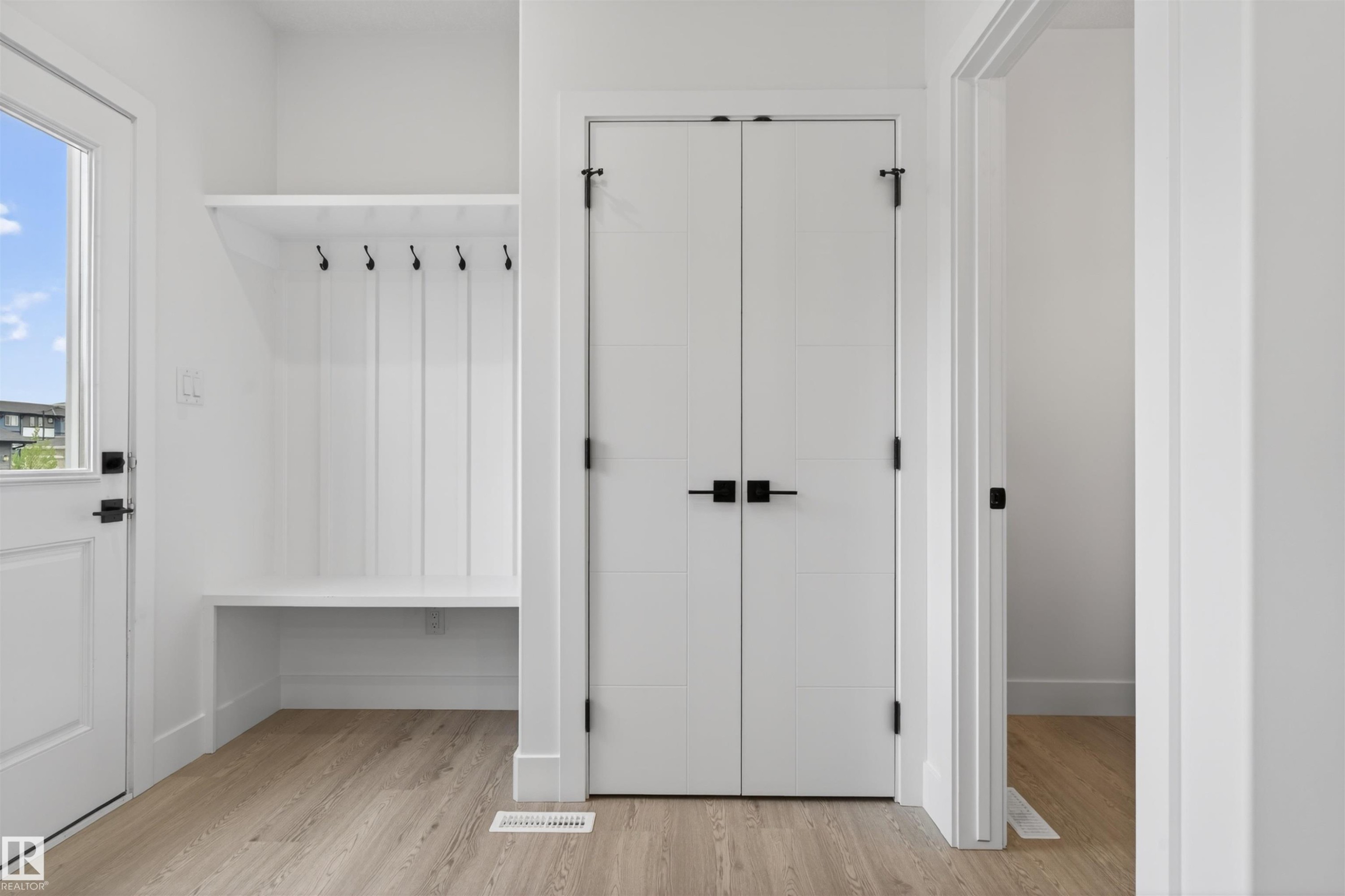 Entryway featuring wood-finish flooring, a white built-in bench with shiplap backing, black coat hooks, and white paneled bi-fold doors with matte black hardware - 45 River Birch, St. Albert, AB - Indoor Photo Showing Other Room