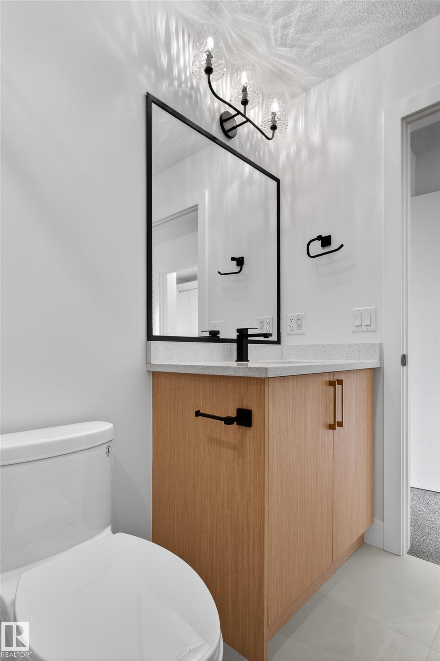 Modern bathroom featuring a wood-finish vanity with a light-colored countertop, a rectangular black-framed mirror, and a three-light sconce with textured glass shades - 45 River Birch, St. Albert, AB - Indoor Photo Showing Bathroom