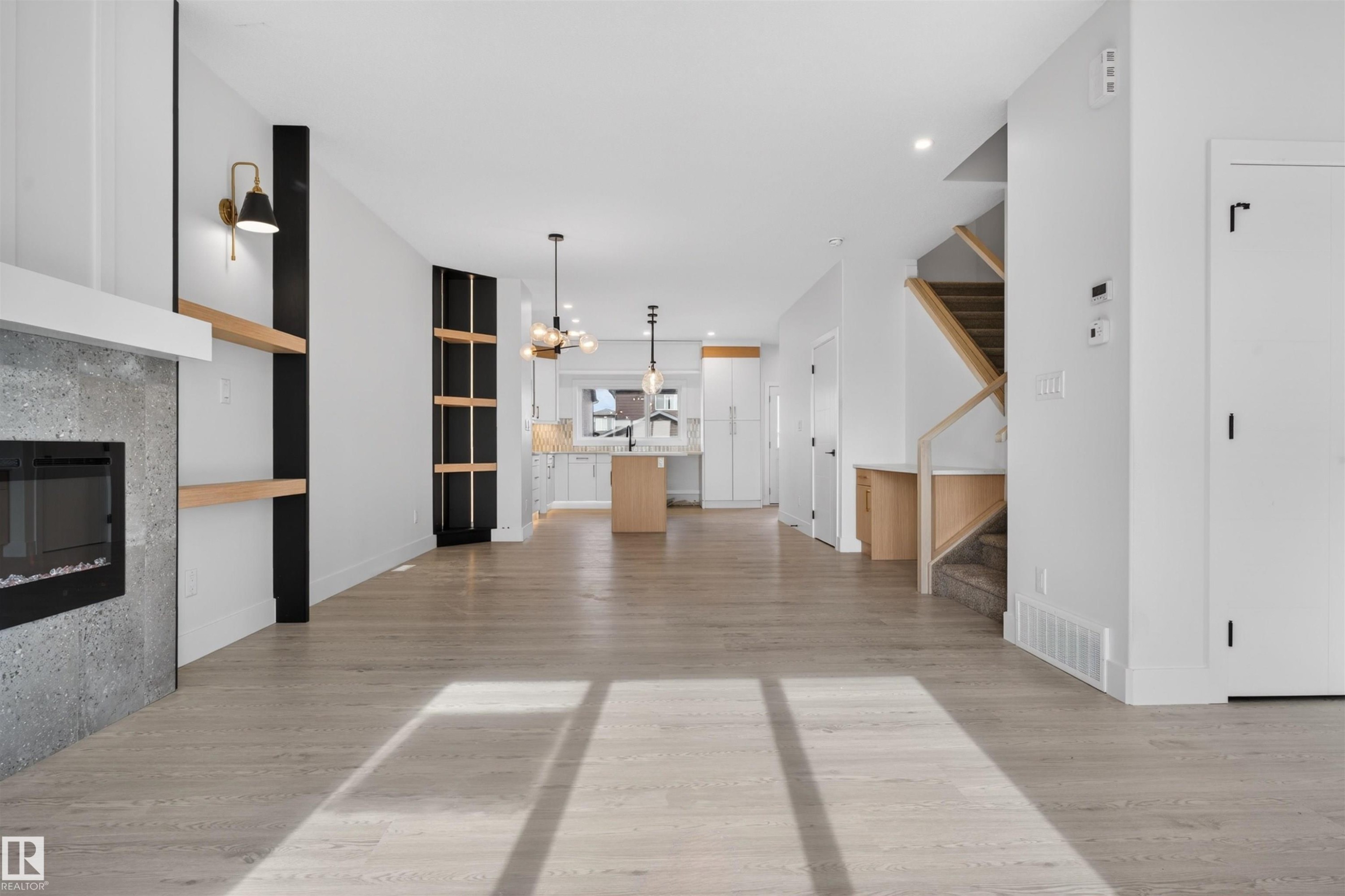 Open-concept living area featuring light wood-finish flooring, a built-in fireplace with a stone-finish surround, integrated shelving with brass sconces, a staircase with a wood-finish railing, and a kitchen with an island and pendant lighting - 45 River Birch, St. Albert, AB - Indoor With Fireplace