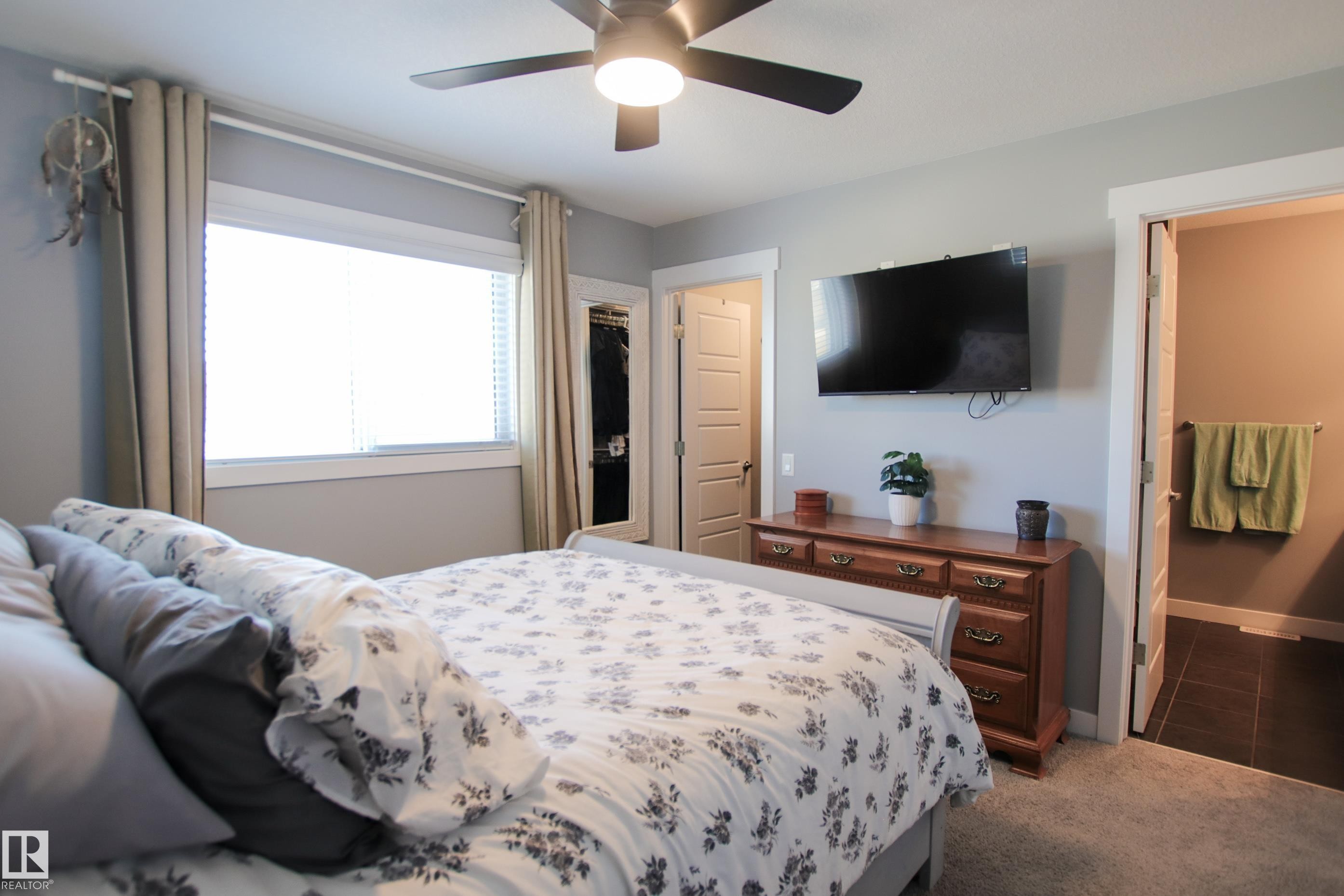 Recessed window with blinds and curtains, ceiling fan with light fixture, carpet flooring, a wardrobe with mirrored door, and a door leading to a bathroom with dark tile flooring - 3805 170A Avenue, Edmonton, AB - Indoor Photo Showing Bedroom