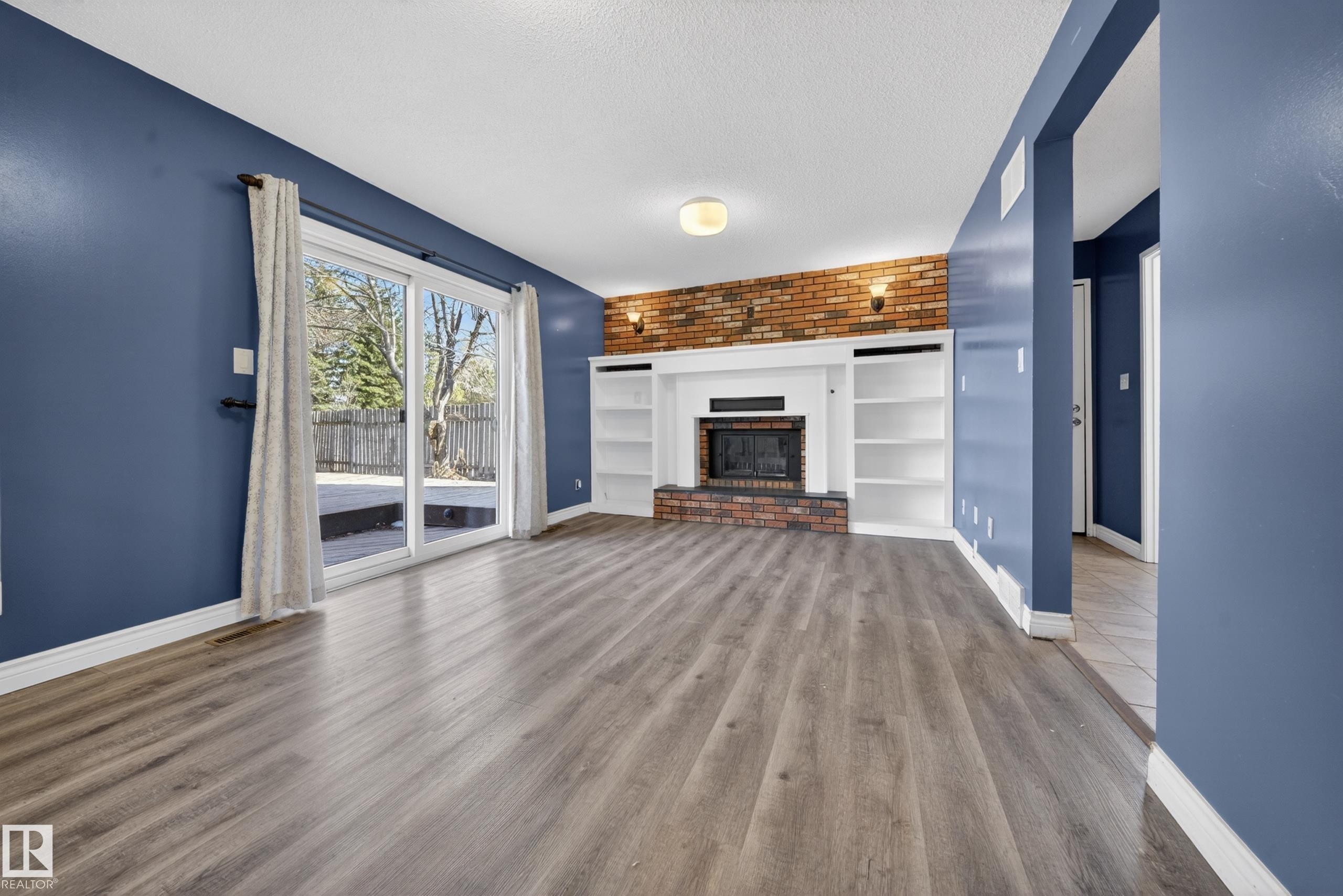 Living area featuring wood-finish flooring, a brick accent wall, built-in shelving, a fireplace with a white mantel, and sliding glass doors leading to an exterior deck - 1767 61 Street, Edmonton, AB - Indoor Photo Showing Living Room With Fireplace