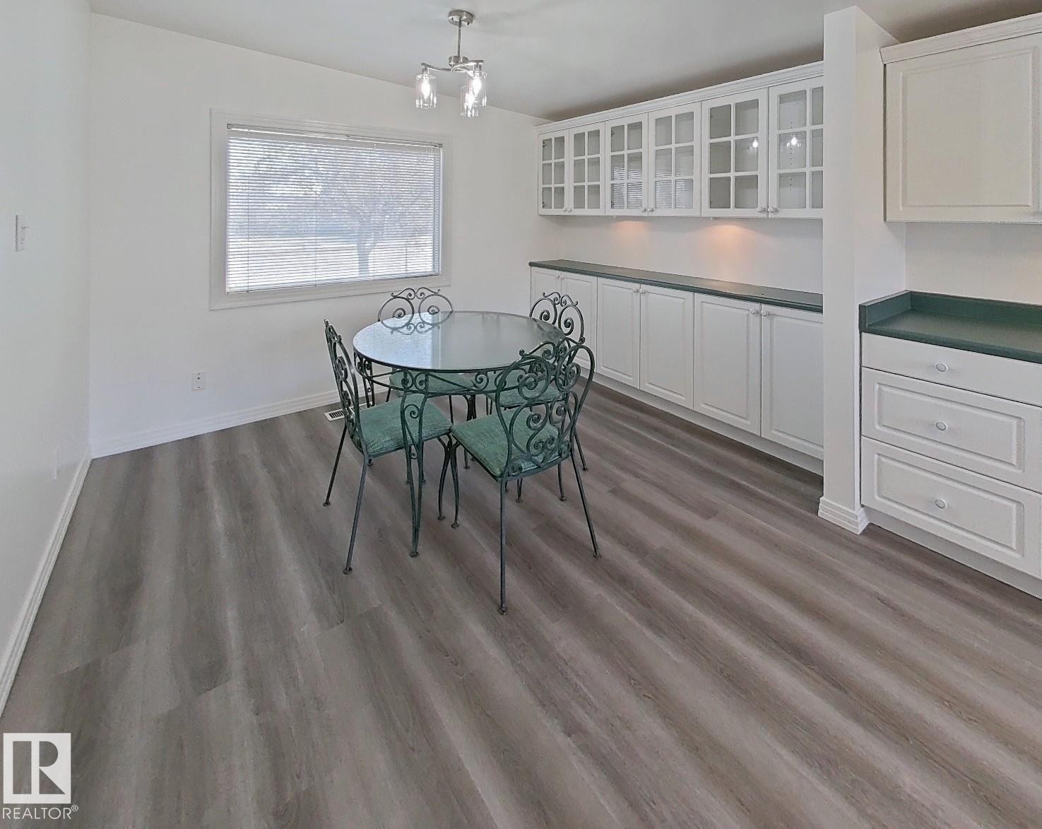 Spacious dining area featuring wood-finish flooring, a large window with blinds, and white built-in cabinetry with glass-front uppers and a dark countertop - 9624 75 Street, Edmonton, AB - Indoor Photo Showing Dining Room