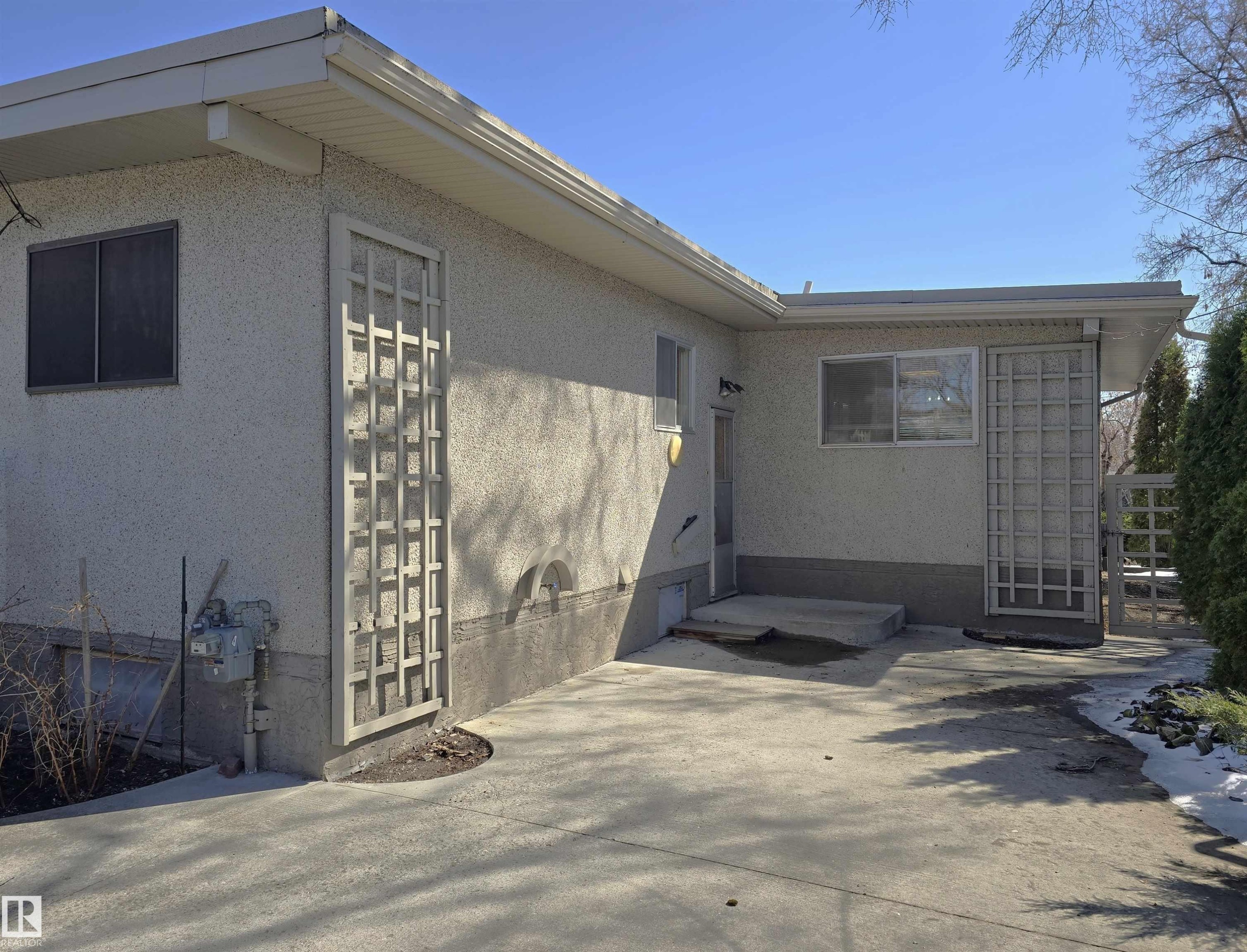 Exterior of a stucco-finished property featuring a concrete patio, decorative lattice panels, and mature evergreens - 9624 75 Street, Edmonton, AB - Outdoor With Exterior