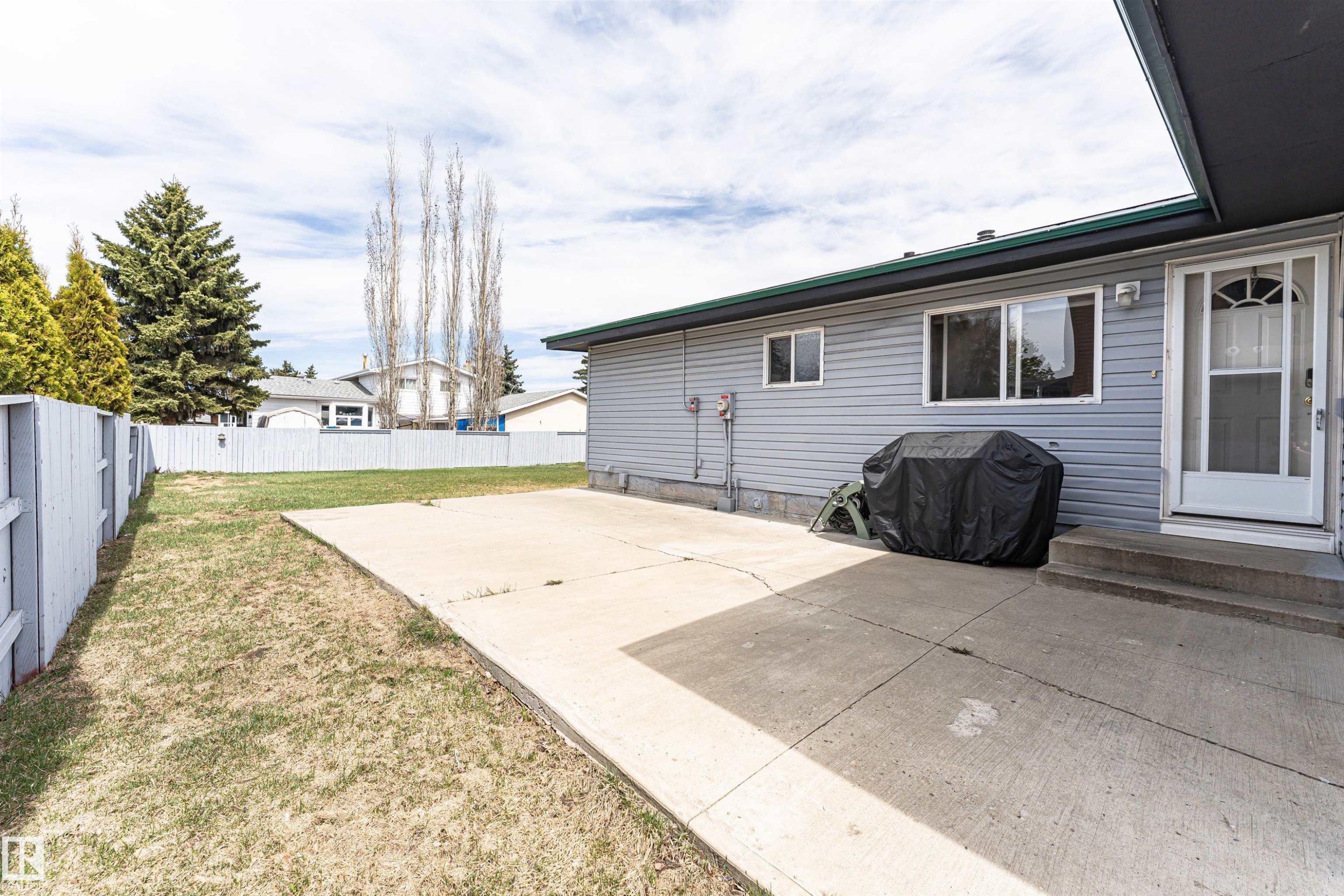 Rear exterior featuring light grey siding, a white storm door, and an expansive concrete patio - 10412 39 Avenue, Edmonton, AB - Outdoor