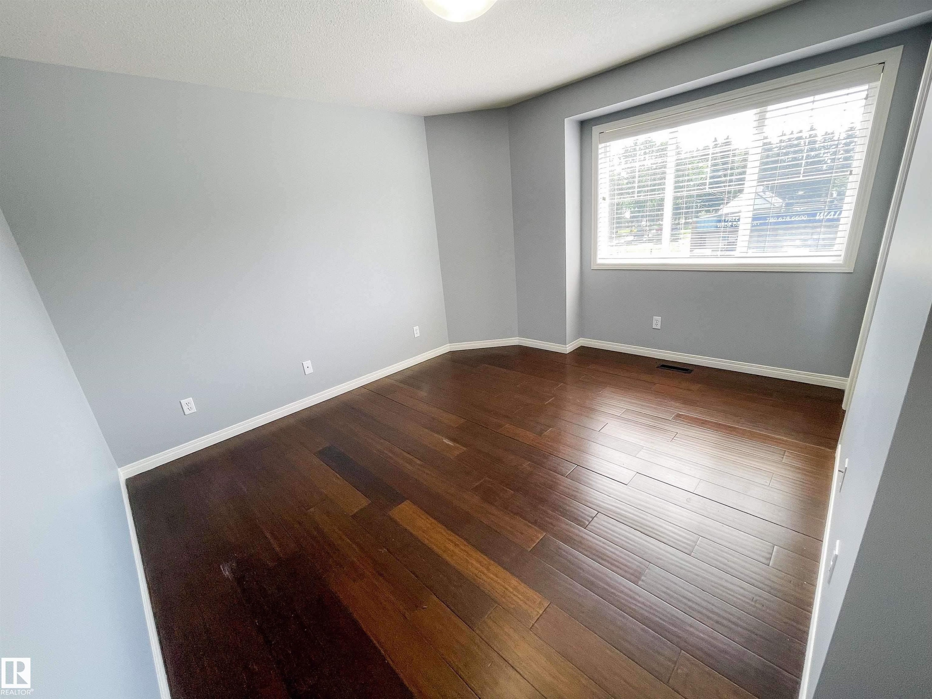 Spacious room featuring rich wood-finish flooring, light grey painted walls, white baseboards, and a large window with white blinds - 5806 118 Avenue Nw, Edmonton, AB - Indoor Photo Showing Other Room