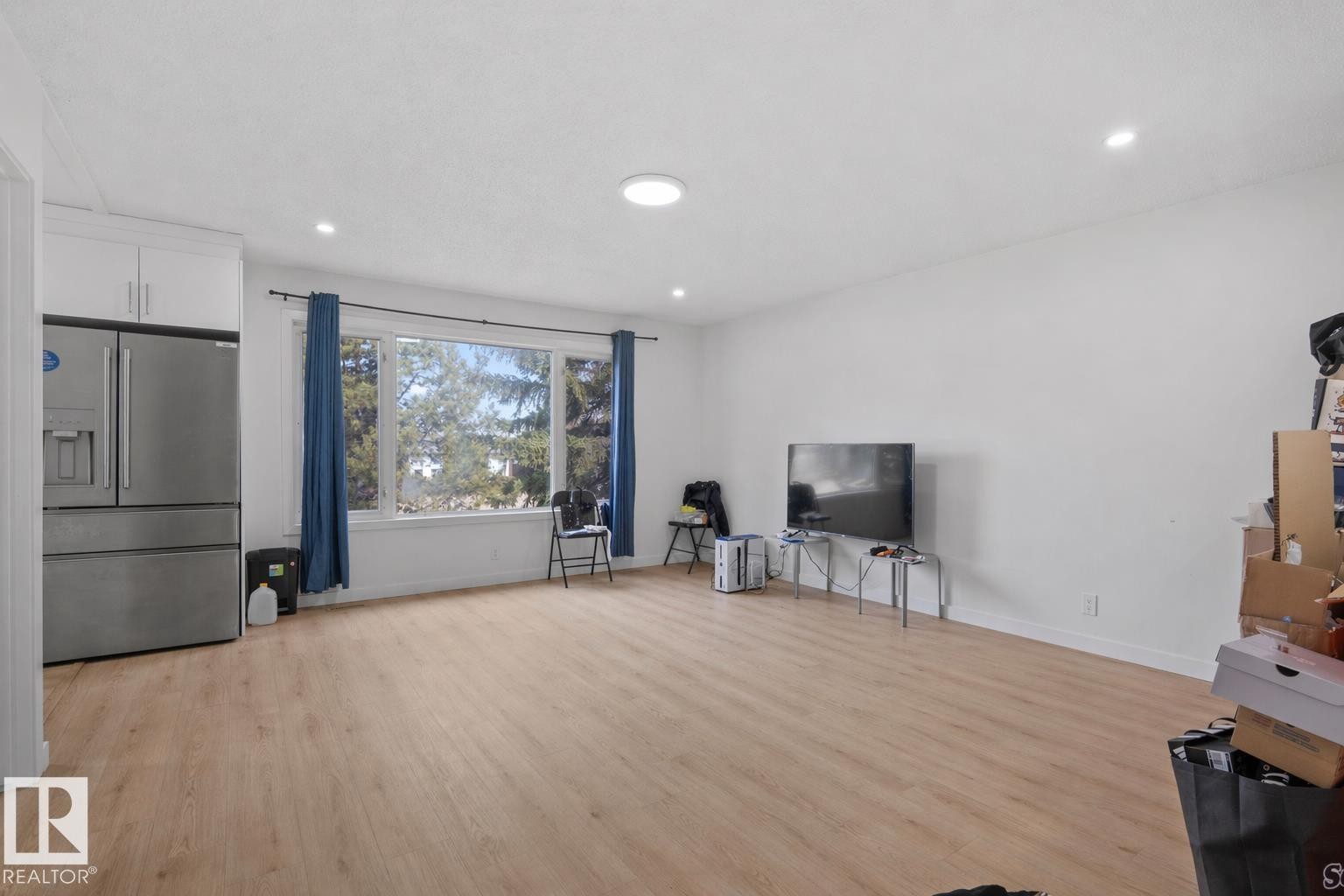 Spacious room featuring light wood-finish flooring, recessed lighting, and a large window with blue drapes - 1566 Lakewood Road Nw, Edmonton, AB - Indoor
