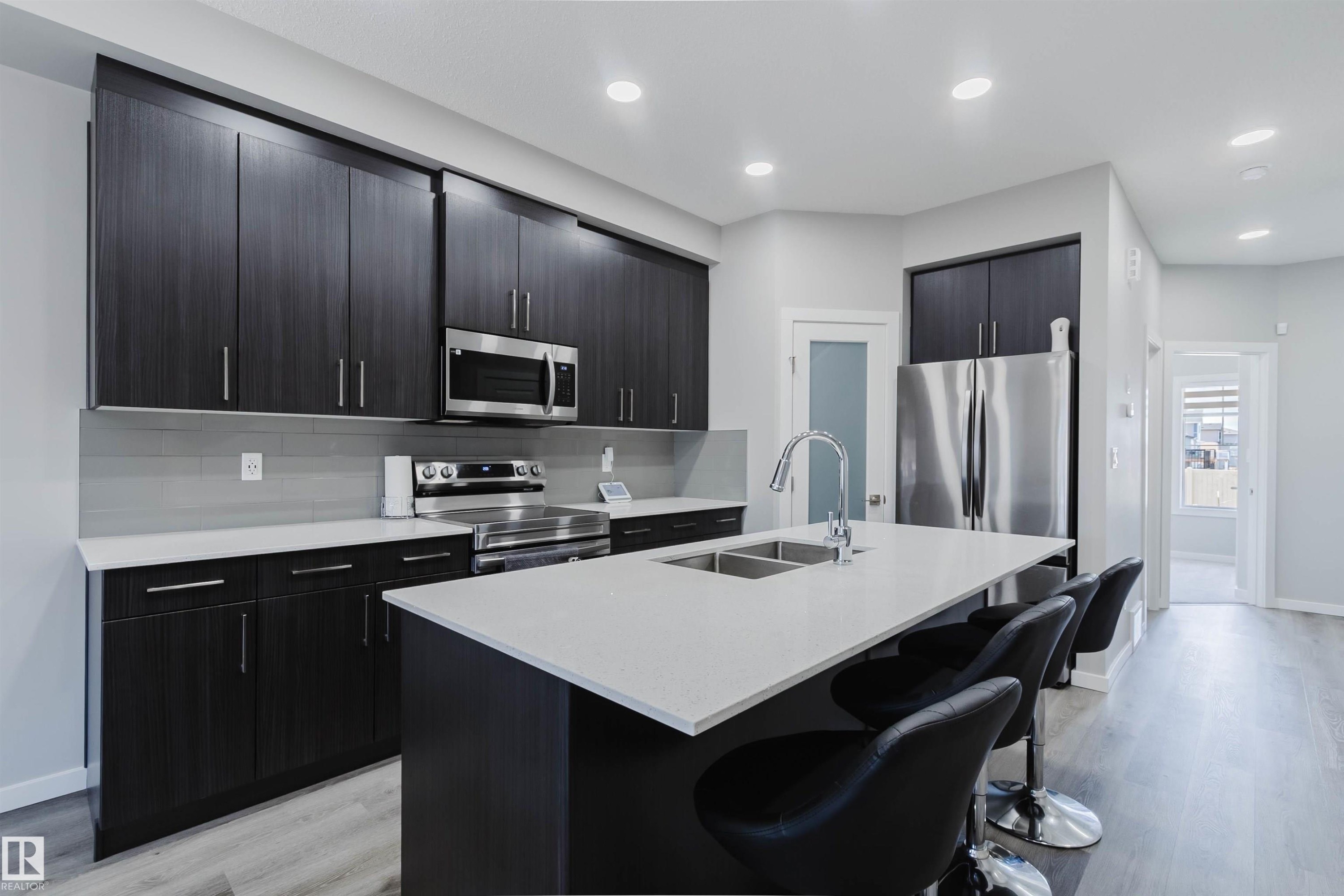 Modern kitchen featuring dark wood-finish cabinetry, stainless steel appliances, a light-toned countertop island with an undermount double sink, and wood-finish flooring - 343 Edgemont Drive, Edmonton, AB - Indoor Photo Showing Kitchen With Stainless Steel Kitchen With Double Sink With Upgraded Kitchen