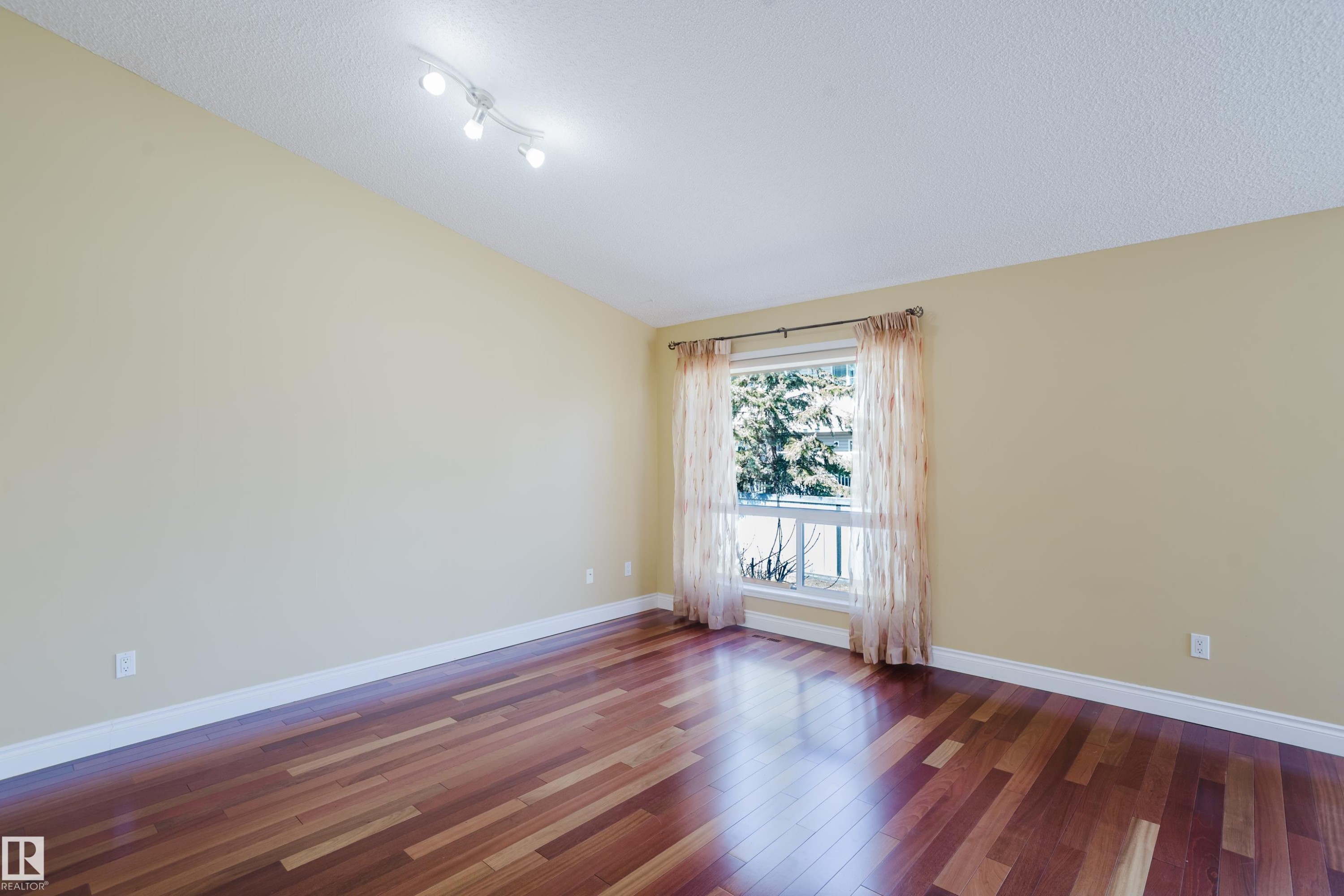 Spacious room featuring wood-finish flooring, a large window, light-toned walls, and a track lighting fixture - 9 102 Bridgeport Crossing, Leduc, AB - Indoor Photo Showing Other Room
