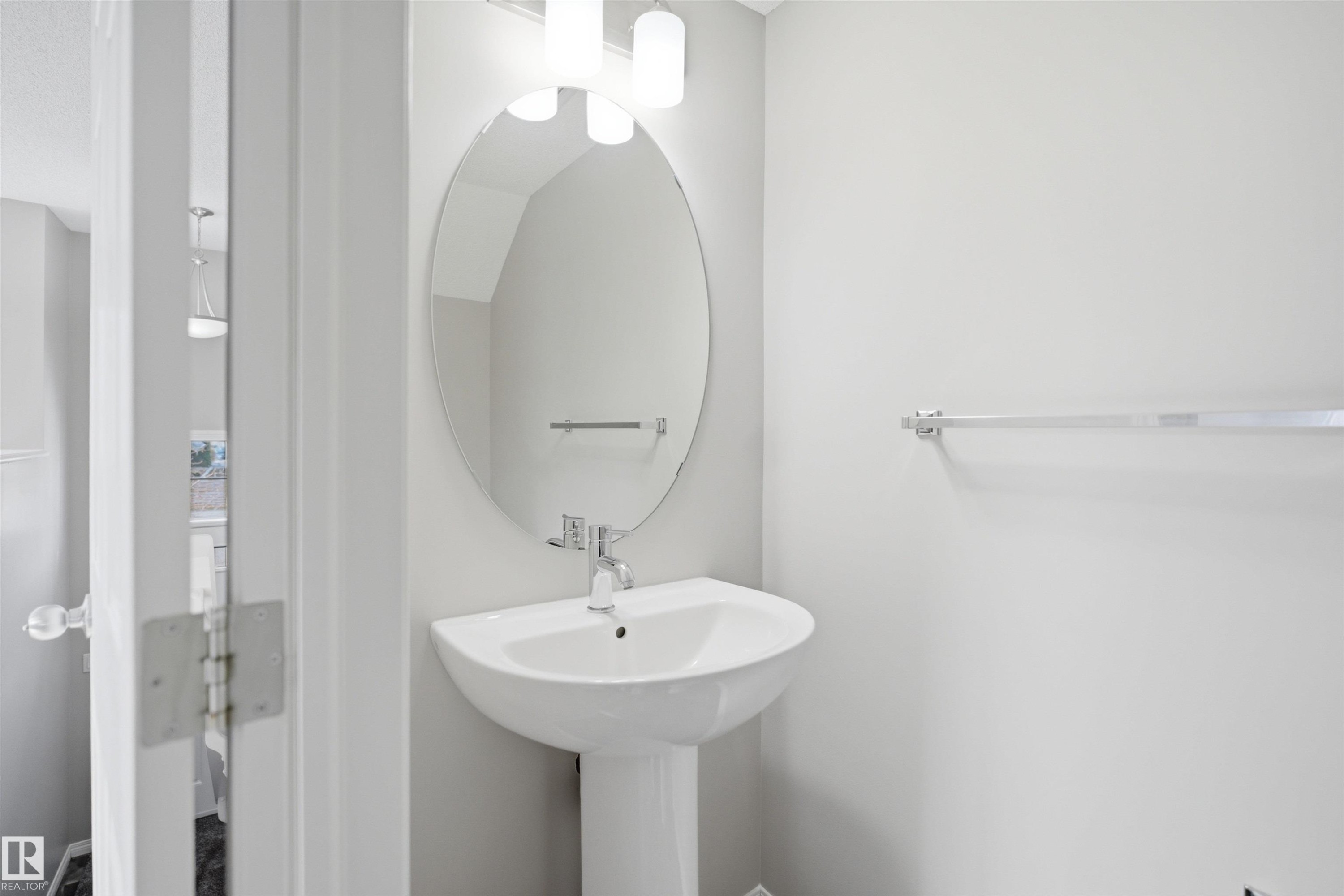 Pedestal sink with chrome faucet, an oval mirror, and wall-mounted light fixture - 33 7385 Edgemont Way Nw, Edmonton, AB - Indoor Photo Showing Bathroom