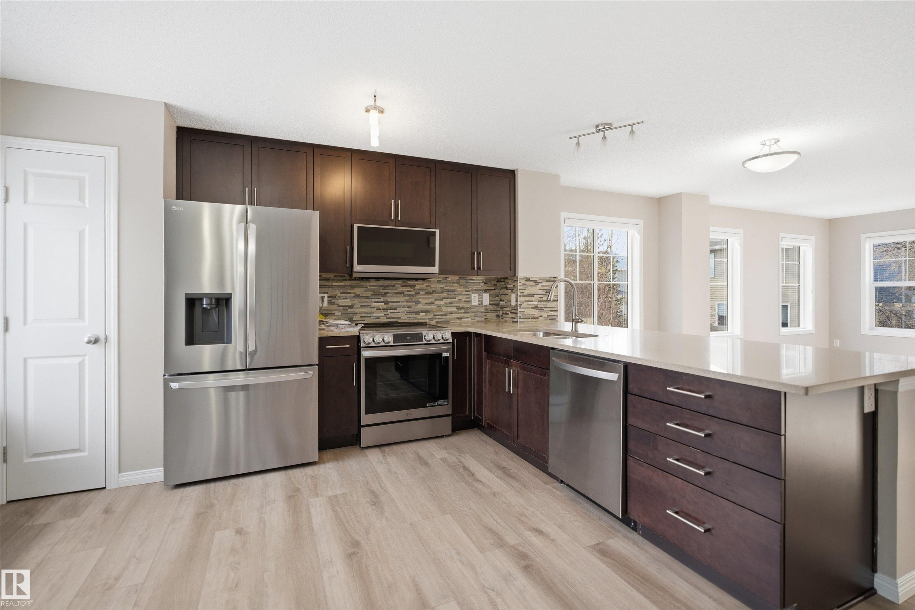 Kitchen featuring dark wood cabinetry, stainless steel appliances, a tiled backsplash, wood-finish flooring, and a large central island with a light-toned countertop - 33 7385 Edgemont Way Nw, Edmonton, AB - Indoor Photo Showing Kitchen With Upgraded Kitchen