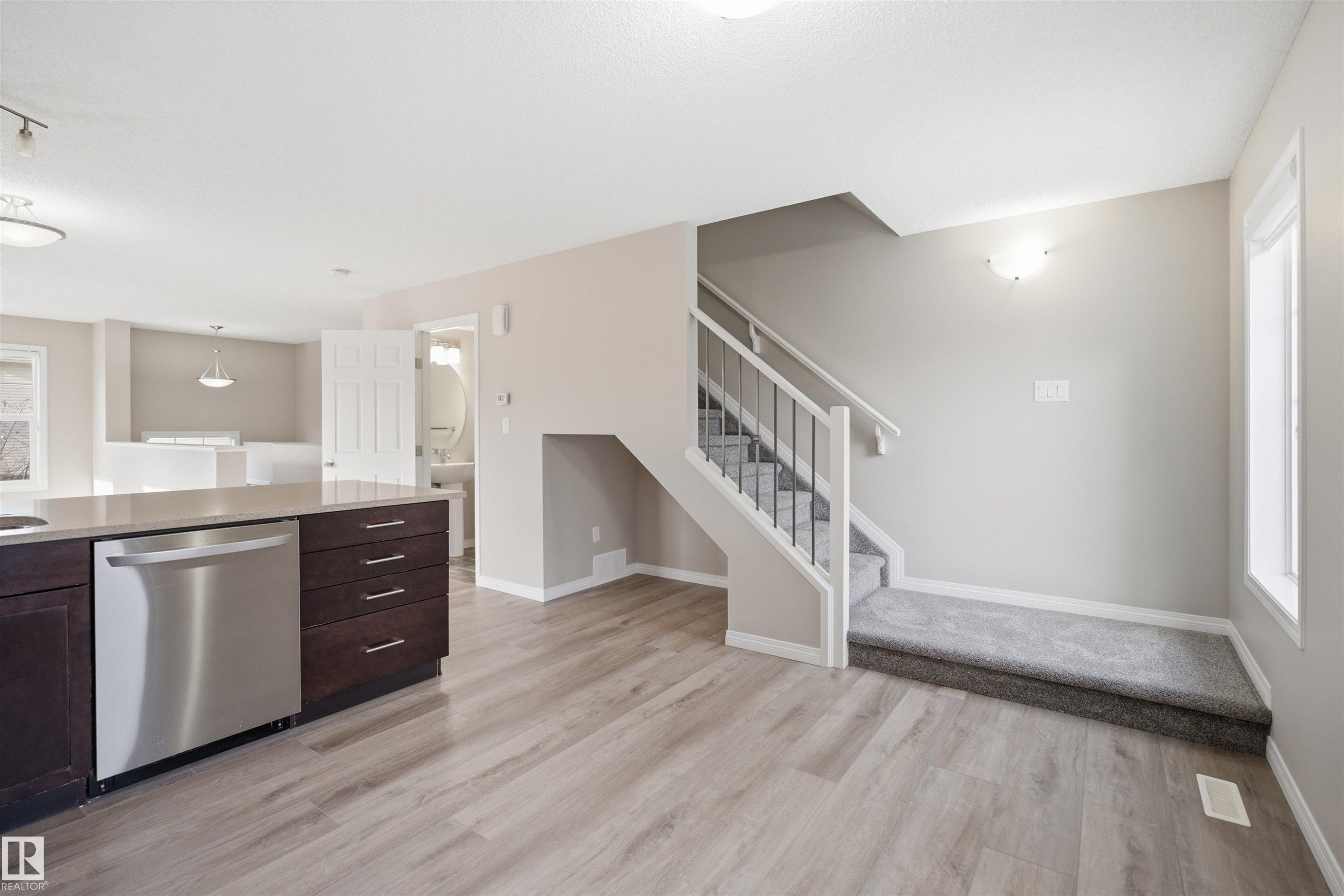 Open-concept living area featuring wood-finish flooring, a kitchen island with stone-finish countertop and stainless steel dishwasher, a carpeted staircase with white risers and black metal spindles, and a recessed wall light fixture - 33 7385 Edgemont Way Nw, Edmonton, AB - Indoor Photo Showing Kitchen