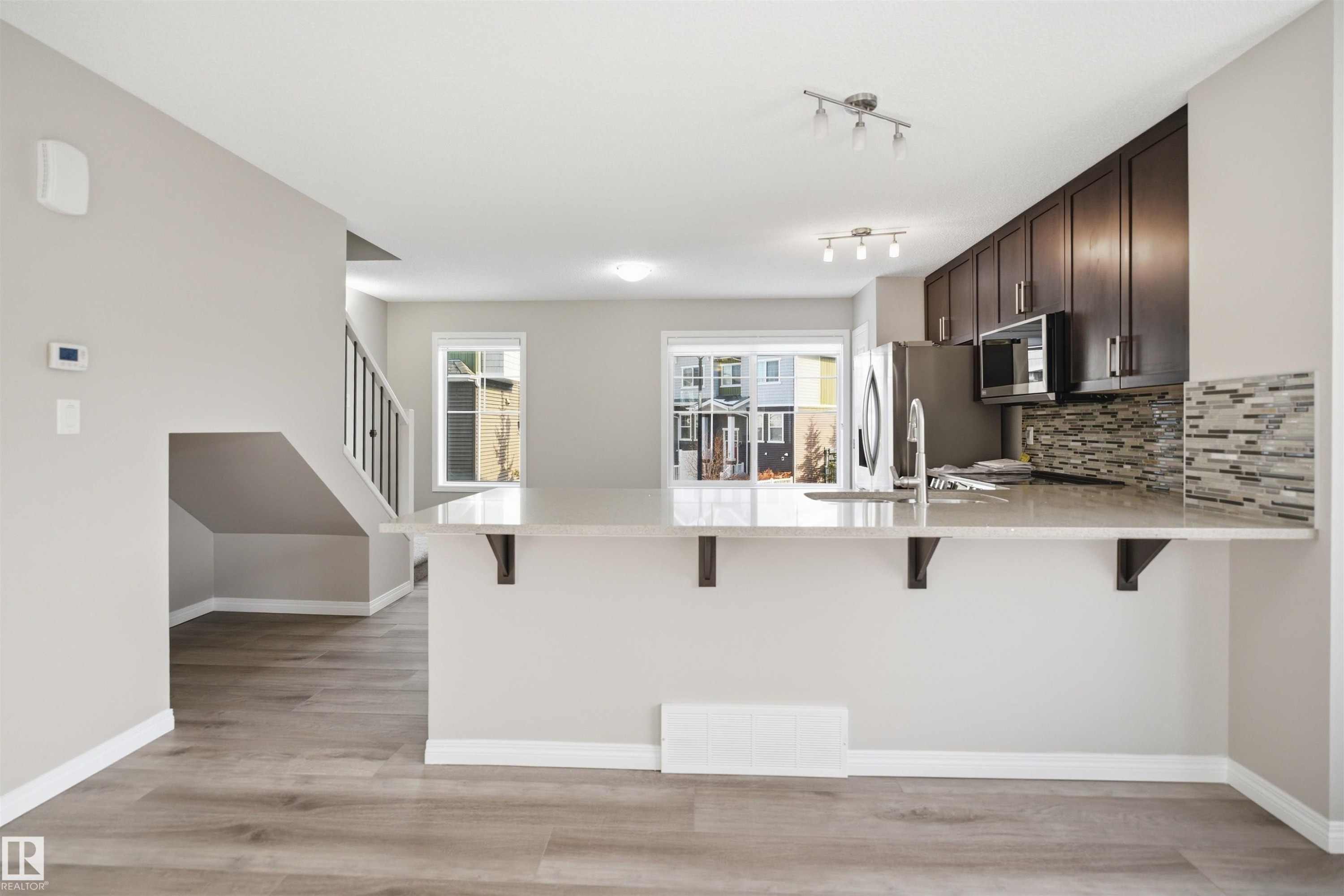 Open-concept kitchen featuring an extended peninsula with light-toned countertop, dark wood-finish cabinetry, stainless steel appliances, and a mosaic tile backsplash - 33 7385 Edgemont Way Nw, Edmonton, AB - Indoor Photo Showing Kitchen With Upgraded Kitchen