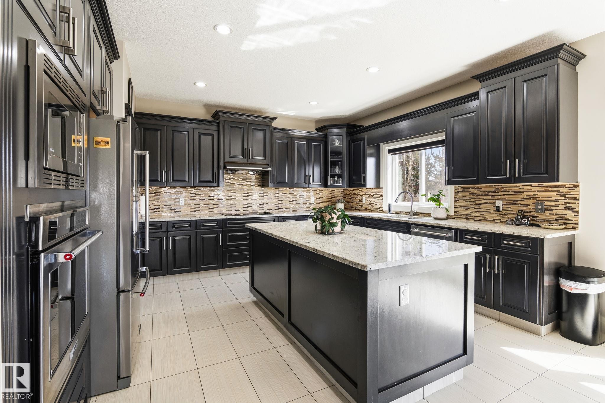 Kitchen featuring dark wood cabinetry, a central island with a light-toned countertop, stainless steel appliances, a mosaic tile backsplash, and light-toned tile flooring - 731 57 Street Sw, Edmonton, AB - Indoor Photo Showing Kitchen With Upgraded Kitchen