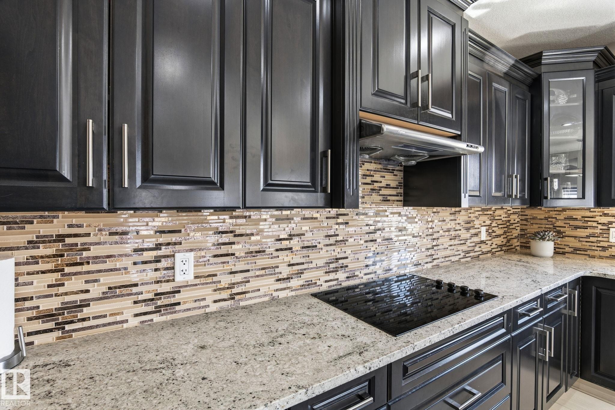 Kitchen featuring dark wood-finish cabinetry with silver-tone hardware, a mosaic tile backsplash, light-colored speckled countertops, and an integrated black cooktop - 731 57 Street Sw, Edmonton, AB - Indoor Photo Showing Kitchen With Upgraded Kitchen