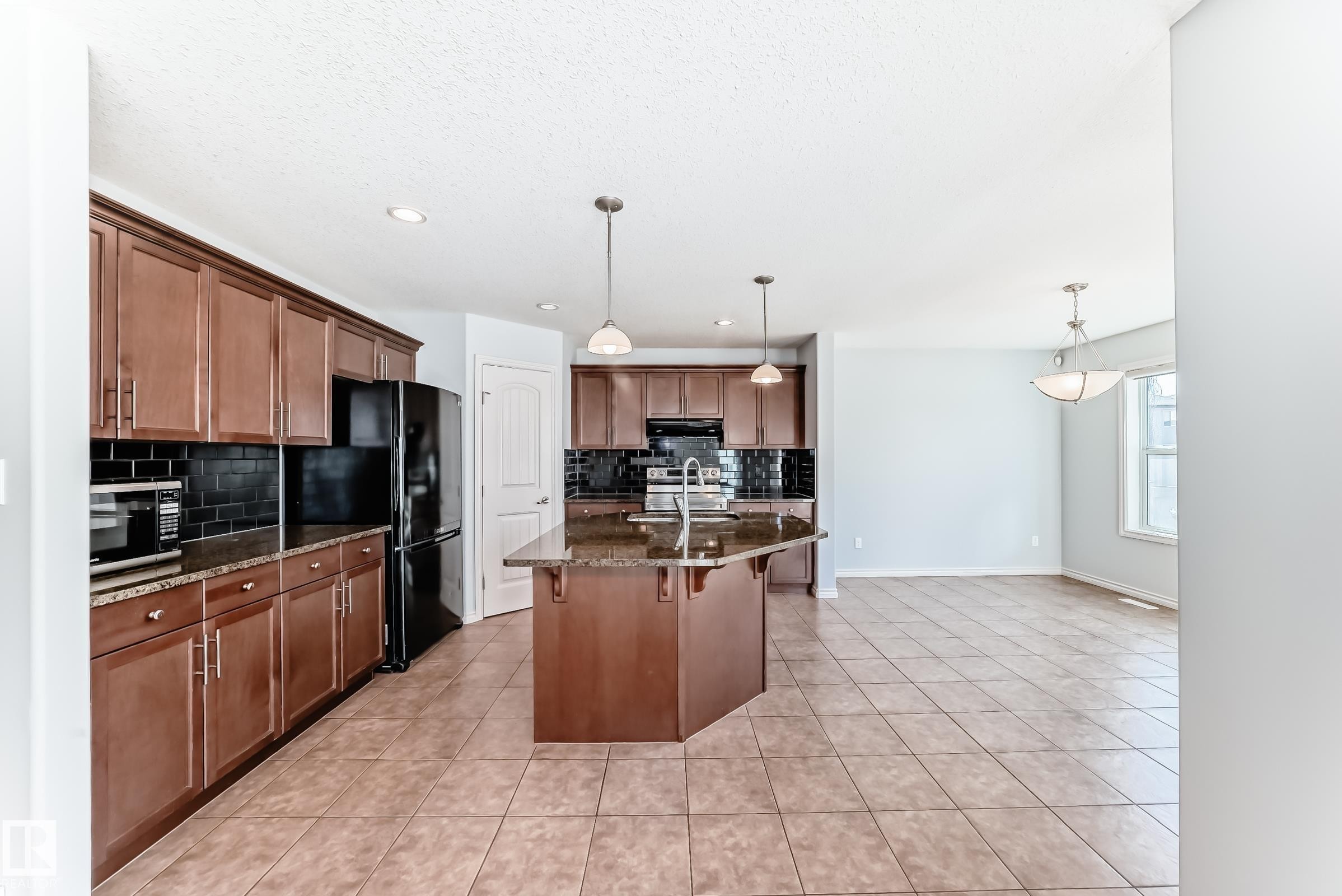 Kitchen featuring an angled island with a double basin sink, stone-finish countertops, dark subway tile backsplash, and coordinating wood-finish cabinetry - 6723 19 Avenue, Edmonton, AB - Indoor Photo Showing Kitchen