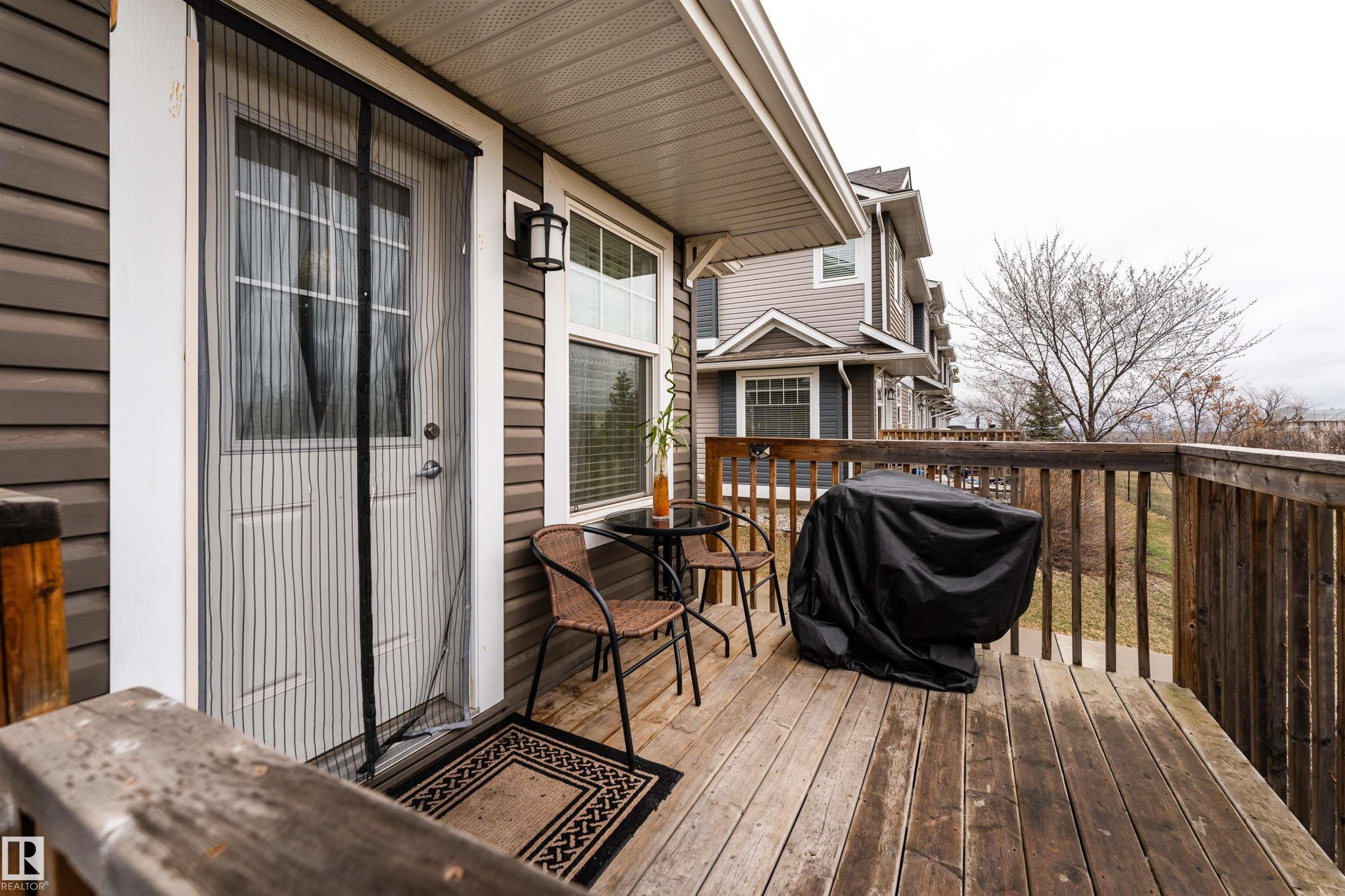 Private outdoor deck featuring horizontal wood siding, a white entry door with glass panels, a window with horizontal blinds, and an exterior wall light fixture - 15 5203 149 Avenue, Edmonton, AB - Outdoor With Deck Patio Veranda With Exterior